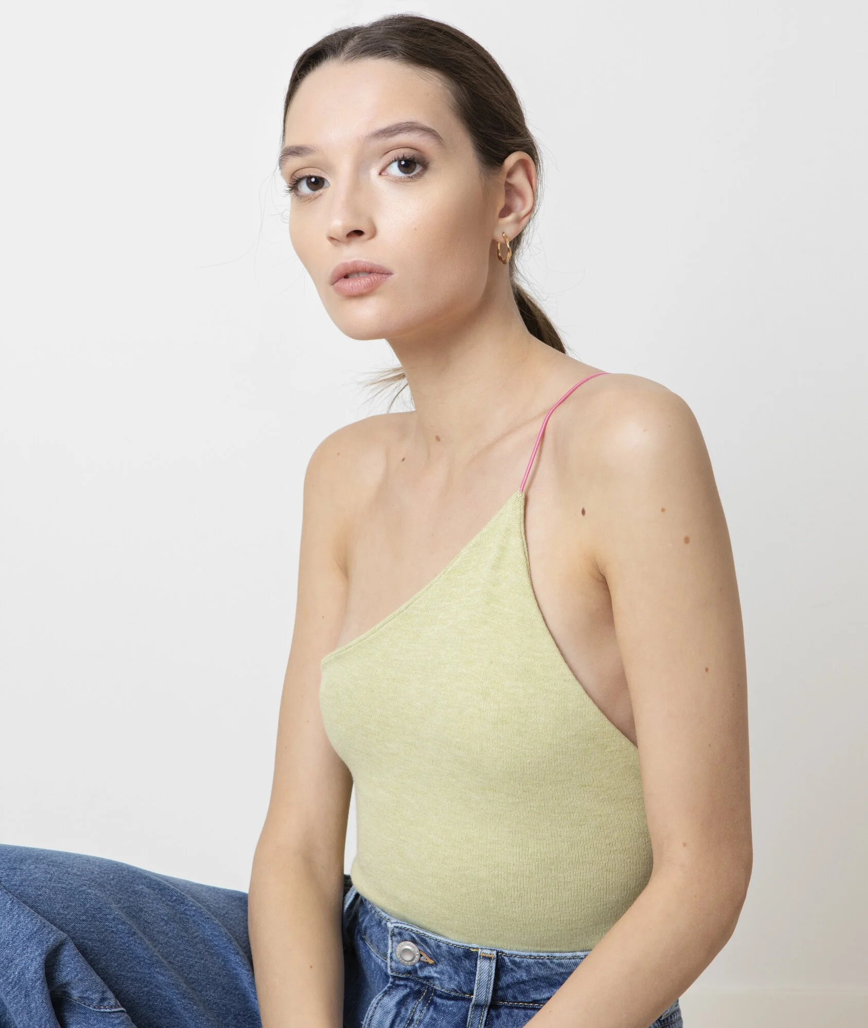 A young woman with brown hair tied back, wearing a light yellow sleeveless top with pink spaghetti straps and high-waisted blue jeans, sitting against a plain white background.