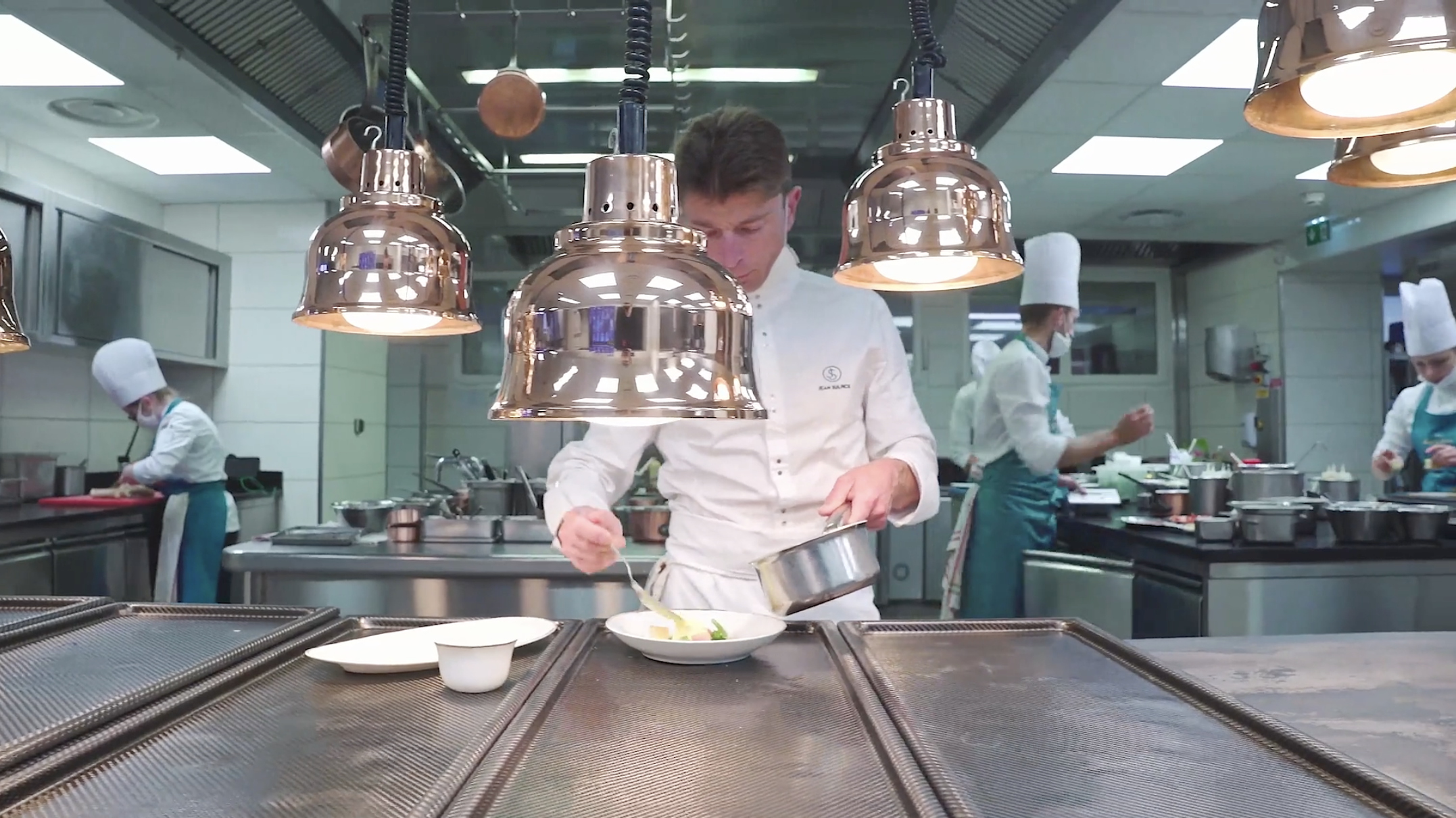 Chef preparing a dish in a professional kitchen with other chefs working in the background.