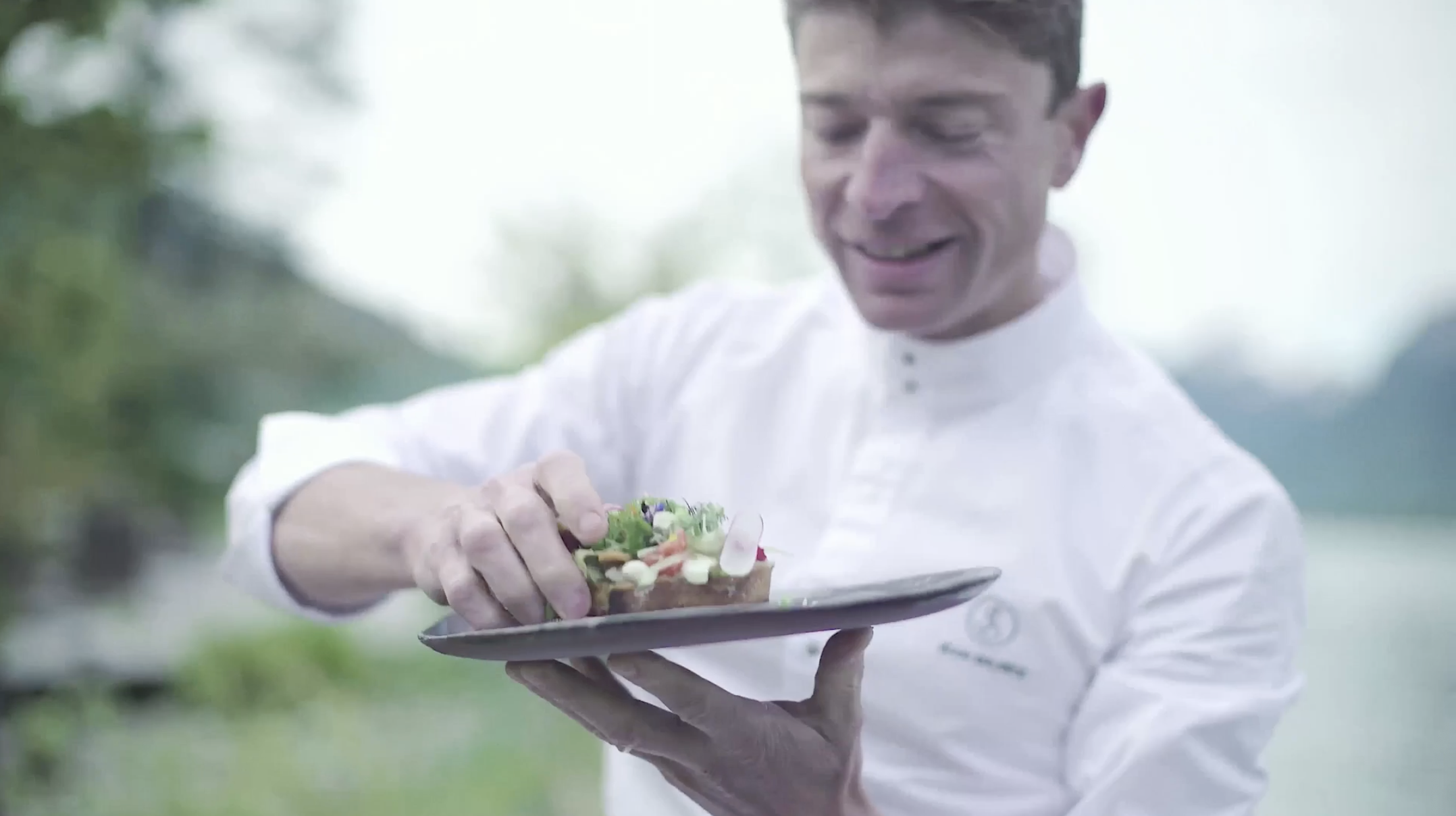 Chef holding a plate with food outdoors, smiling.