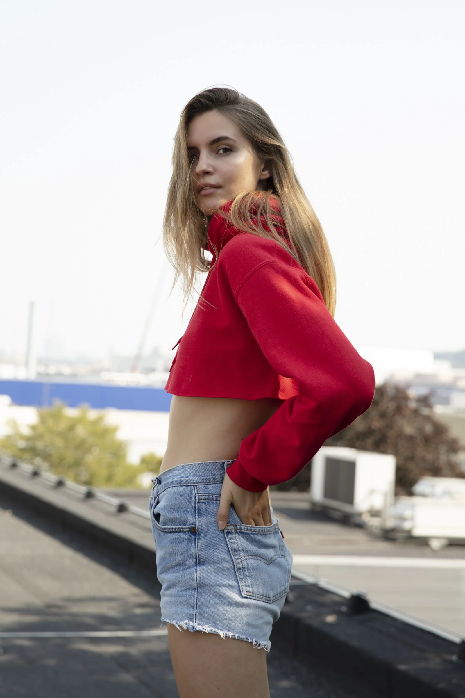 A young woman with long hair wearing a red crop top and denim shorts standing outdoors on a rooftop during the daytime.
