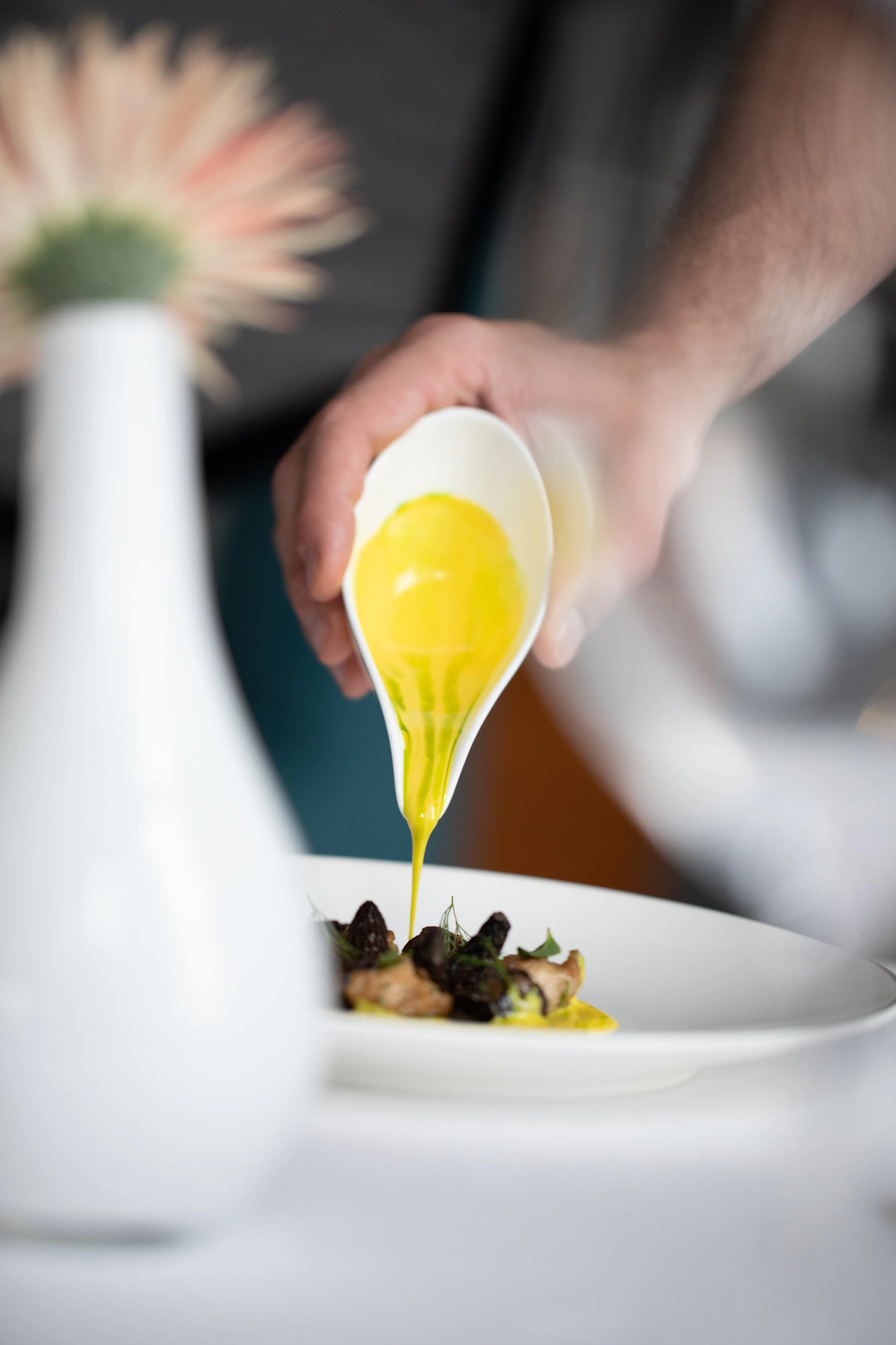 Chef pouring yellow sauce over a plated dish garnished with greens and dark ingredients, with a blurred flower in the foreground.