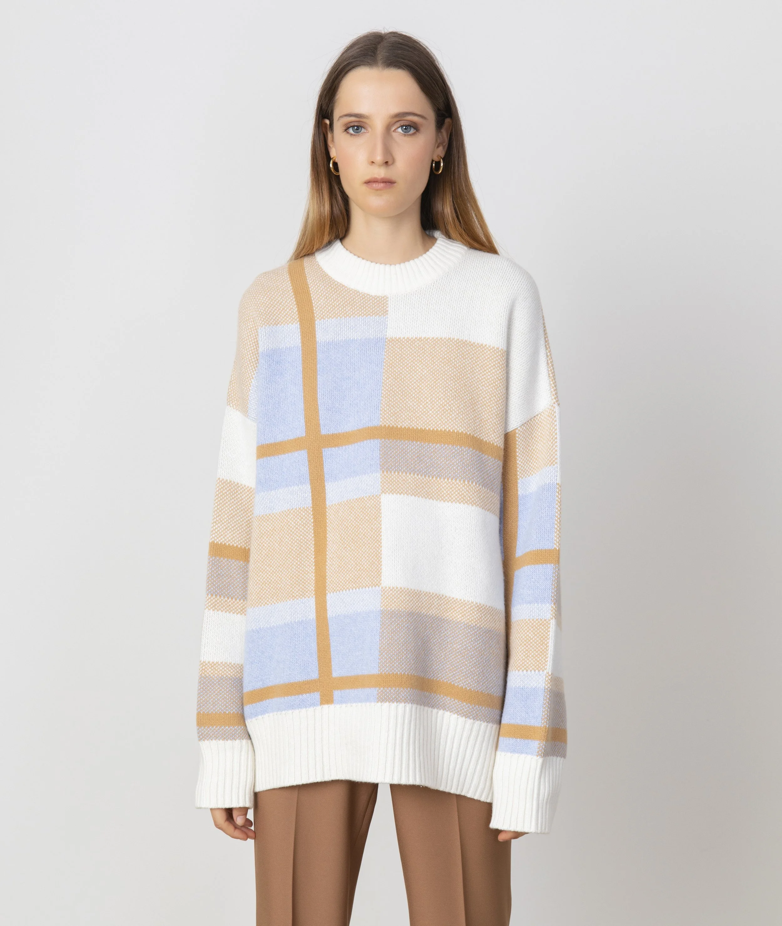 A woman with long brown hair wearing a white, blue, and tan plaid-patterned oversized sweater and tan pants, standing against a plain white background.