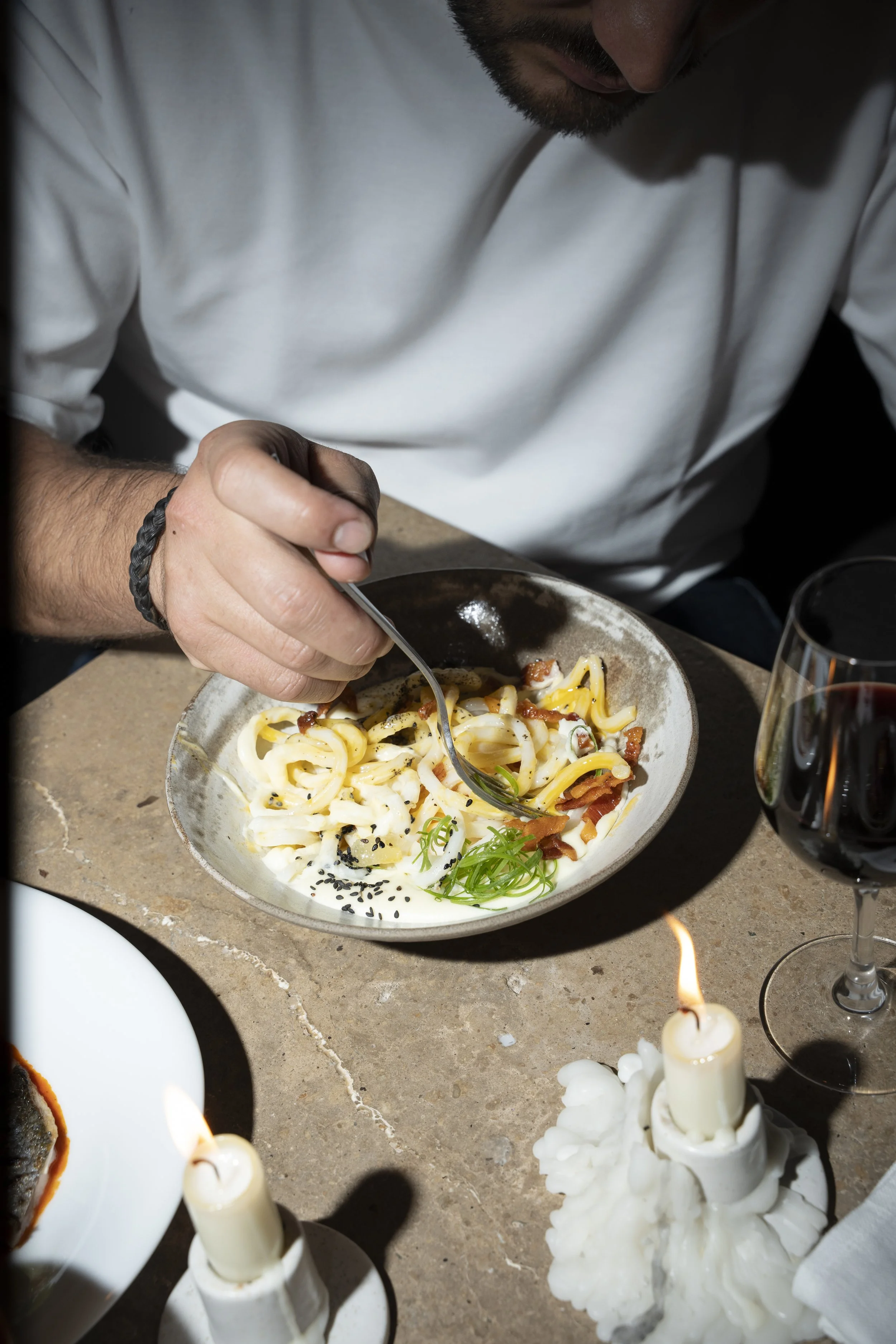 Person eating pasta with black sesame seeds, bacon, and green onions at a table with lit candles and a glass of red wine.