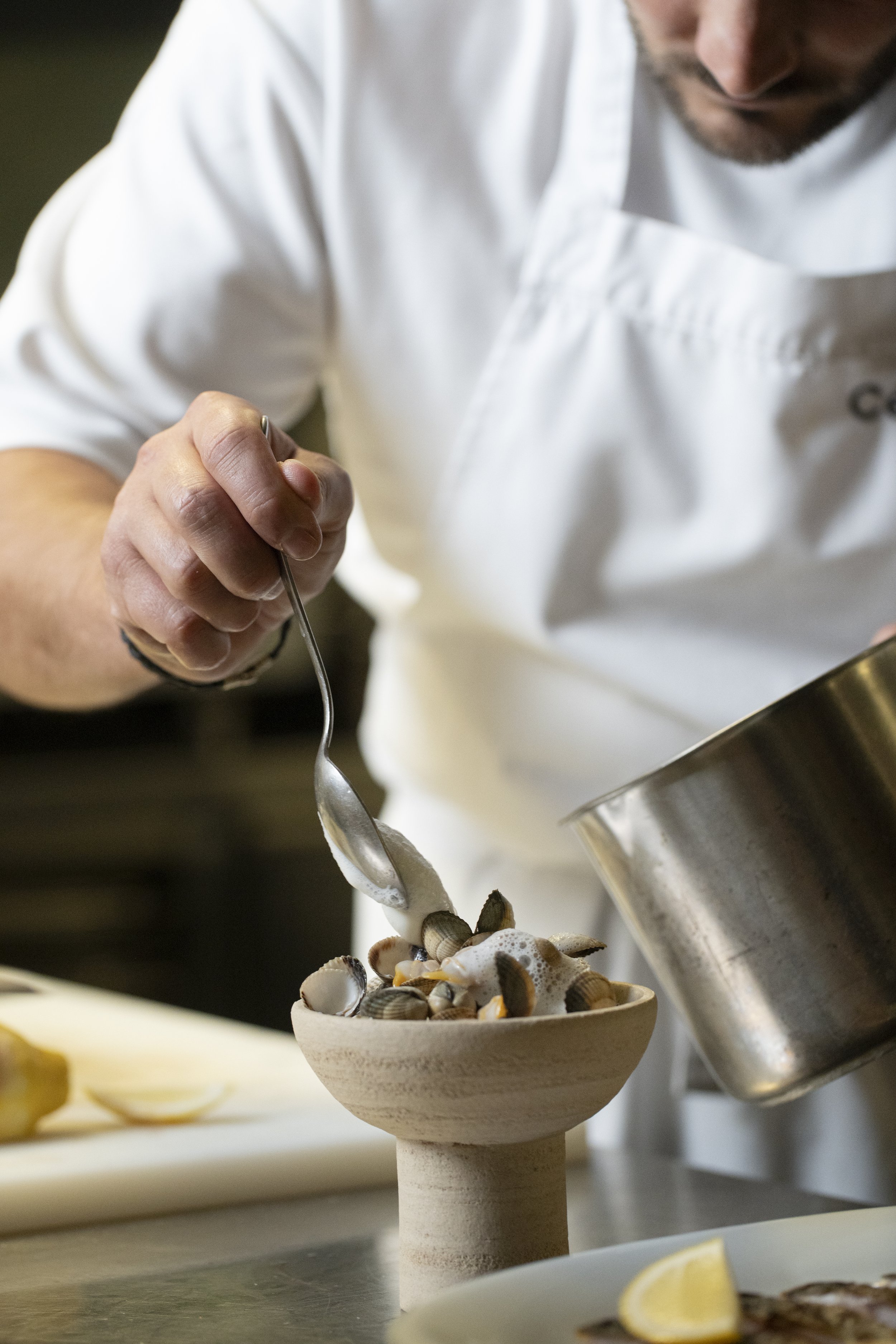 Chef in a white coat preparing a bowl of cooked clams with foam on top, using a spoon in a professional kitchen.