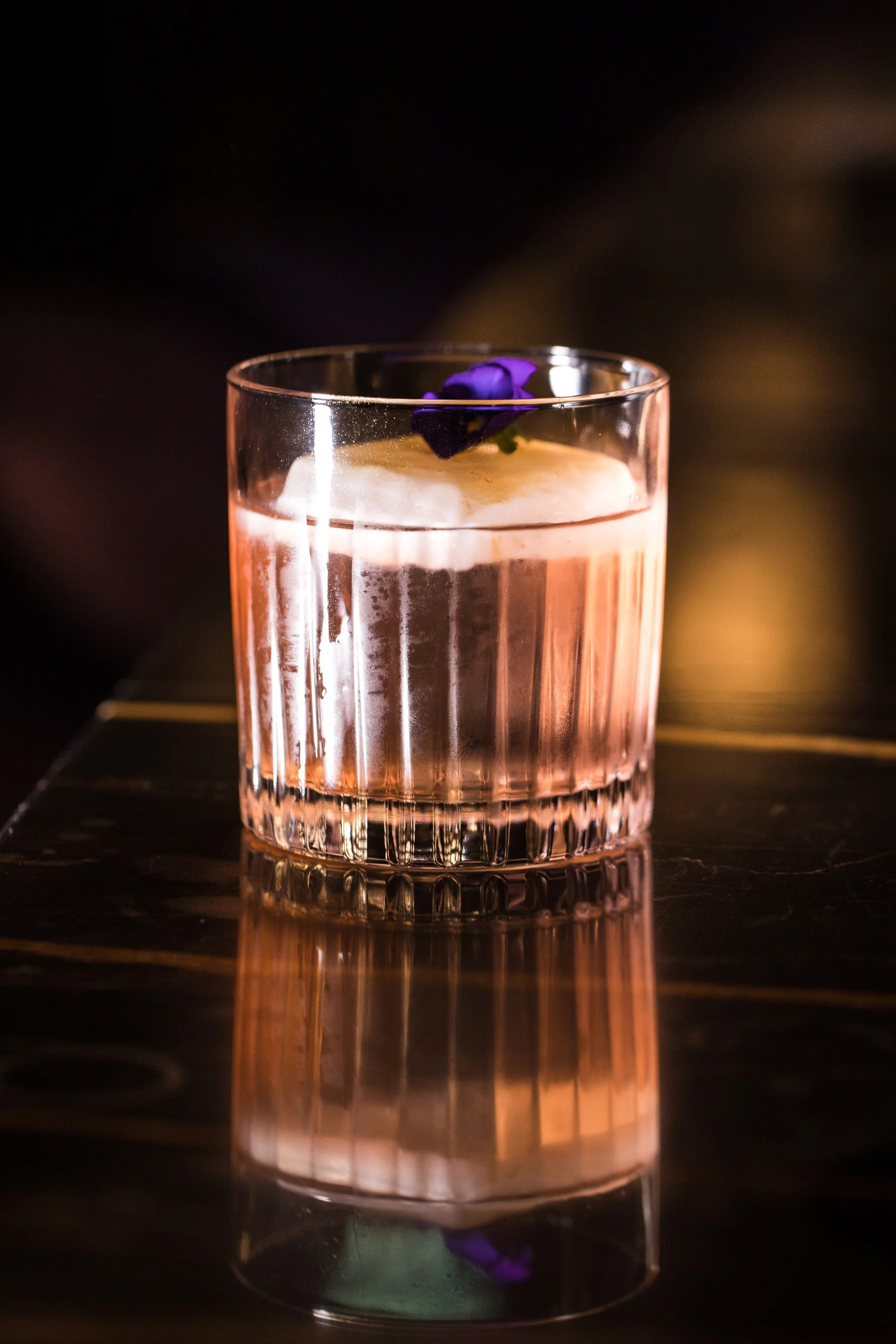 A glass of pink cocktail with a purple flower garnish on top, reflected on a dark surface.