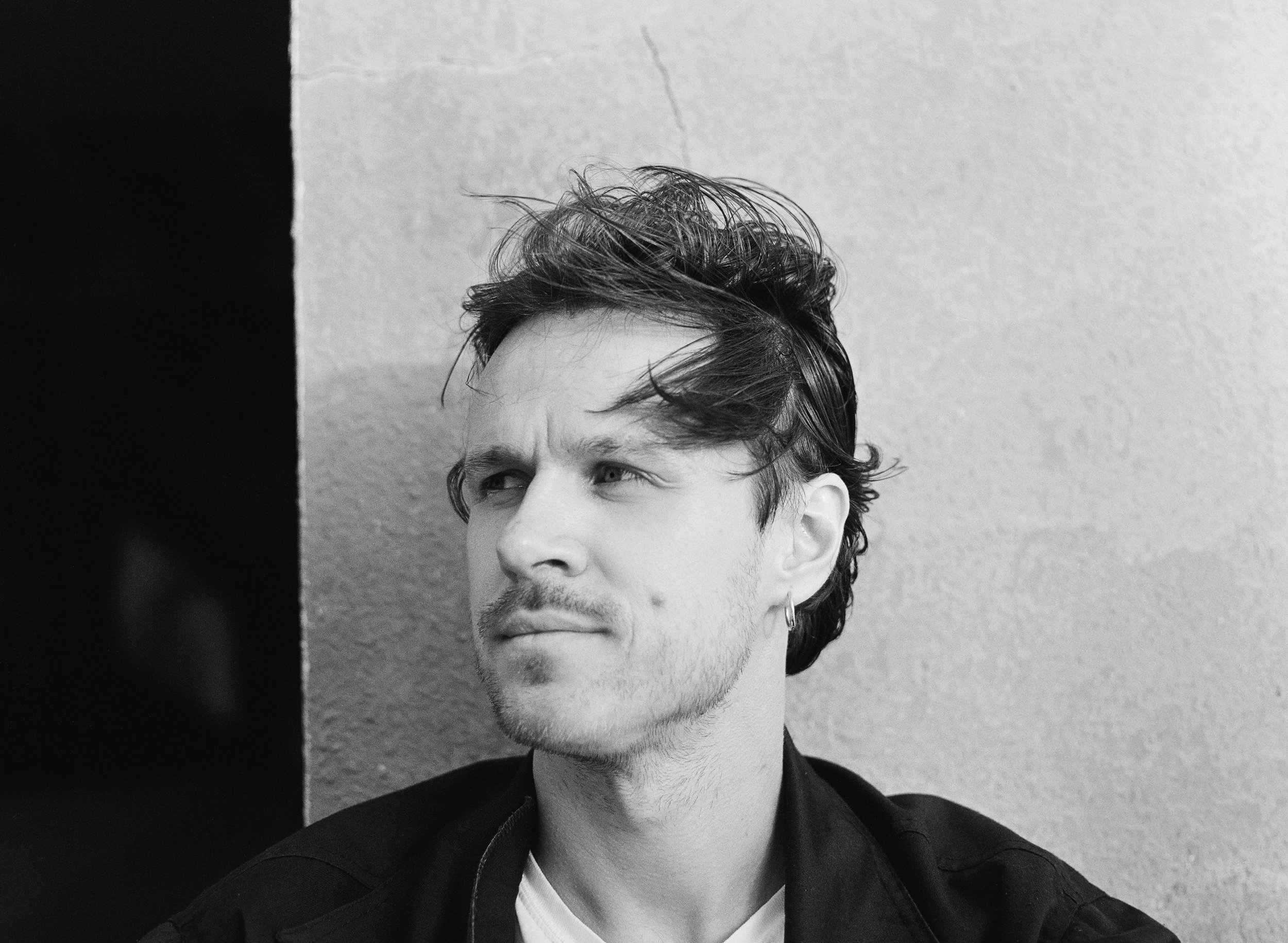 Black and white photo of a young man with messy hair, a small earring, and a slight beard, looking contemplative against a textured wall background.