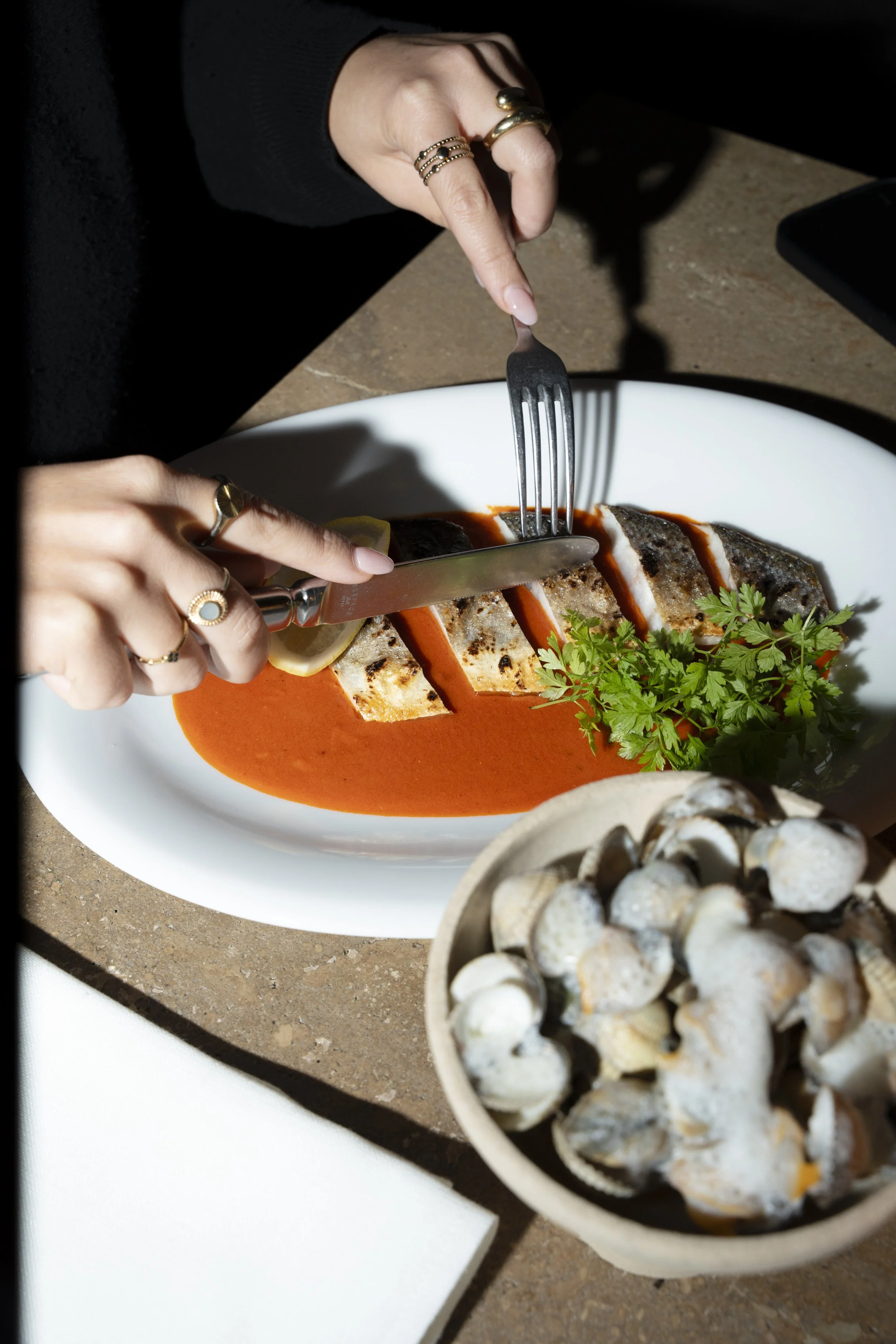 Person slicing grilled fish with lemon in a red sauce on a white plate, garnished with cilantro, with a bowl of cooked shellfish nearby.