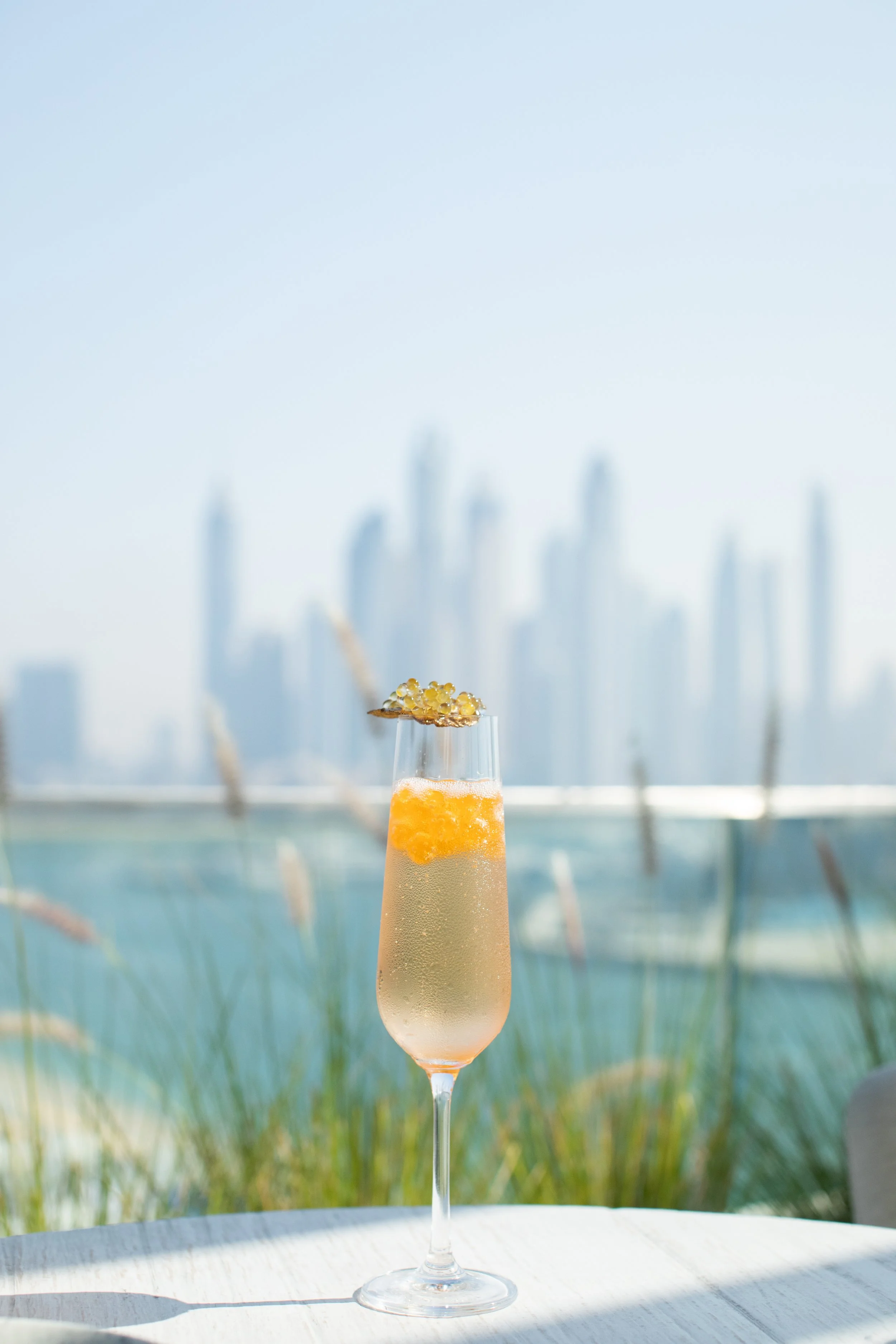 A glass of champagne with a citrus garnish and a decorative topping, set on a white table with a blurred city skyline and waterfront in the background.