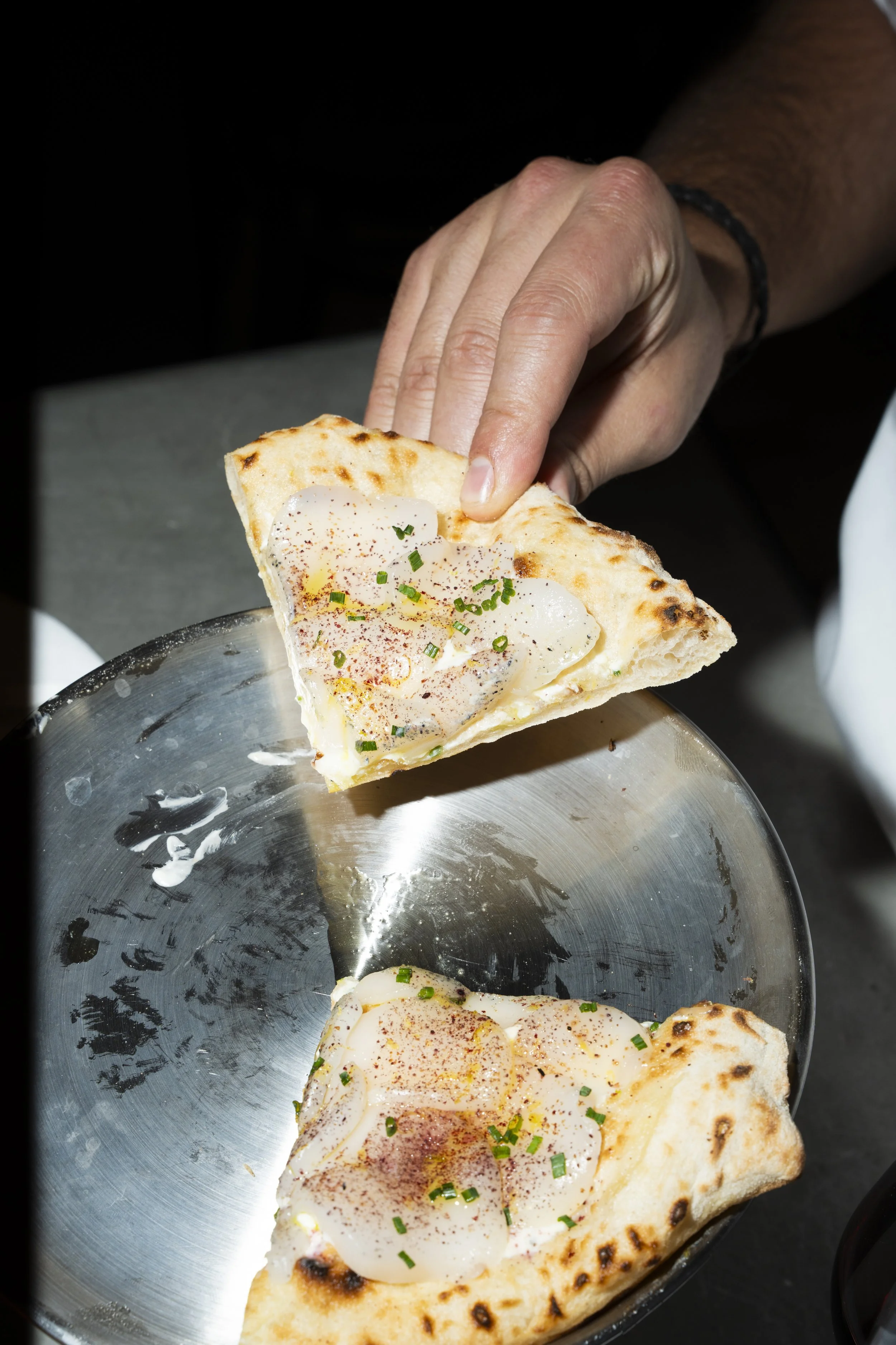 Person holding a slice of pizza with eggs, garnished with green herbs, served on a stainless steel plate.