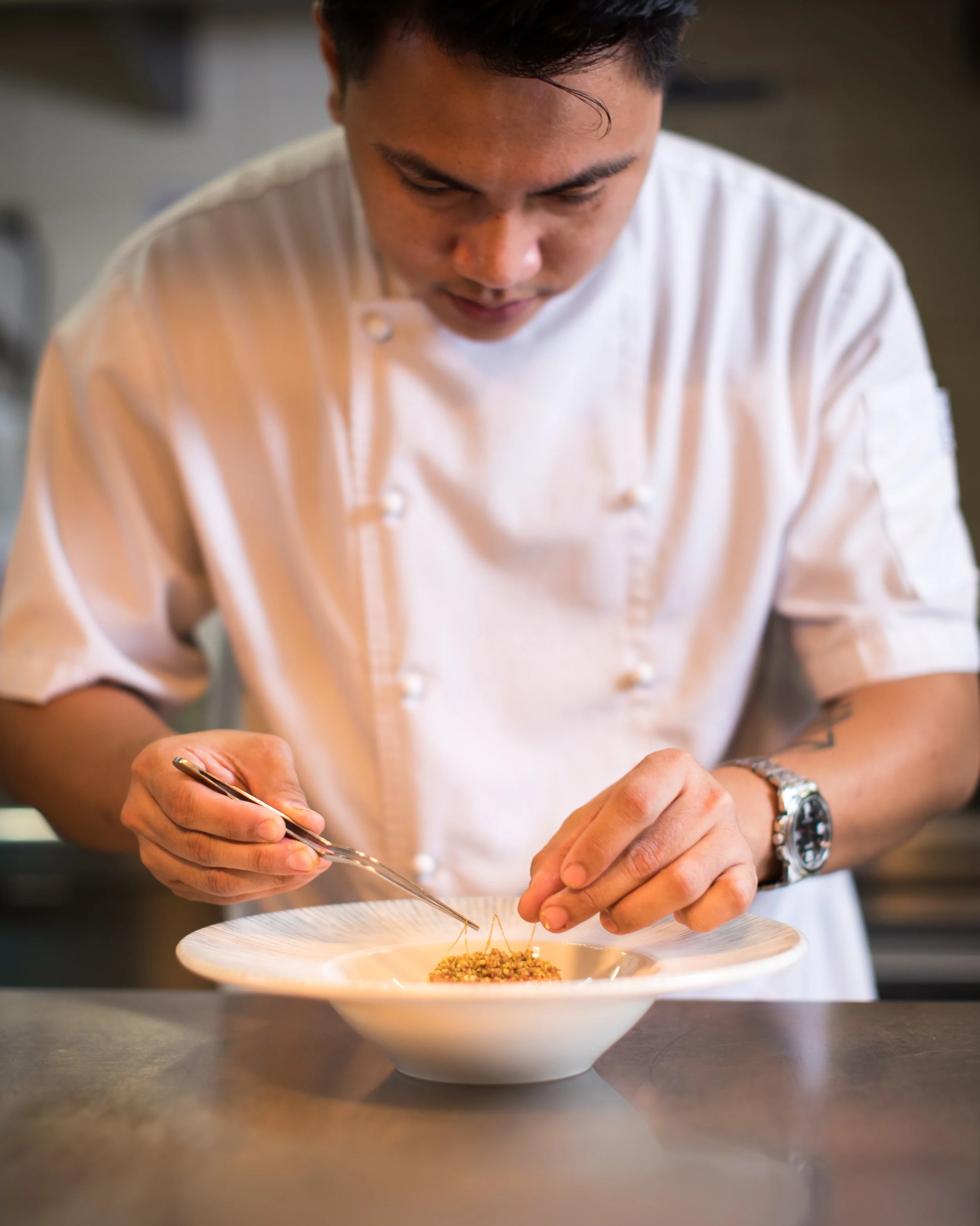 Chef in a white uniform plating a dish with tweezers in a professional kitchen.
