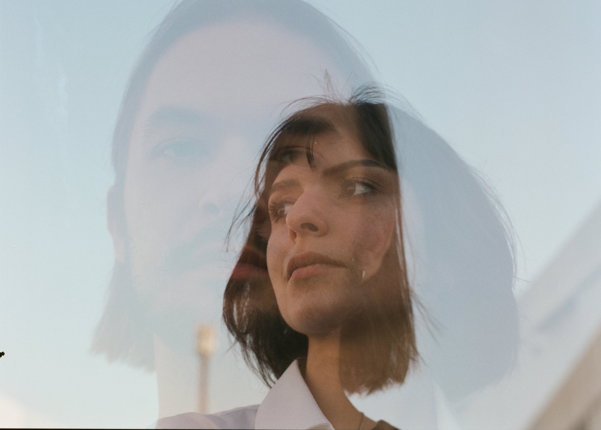 A double exposure photograph of a woman with dark hair and a neutral expression against a pale sky, creating an overlay of her face.