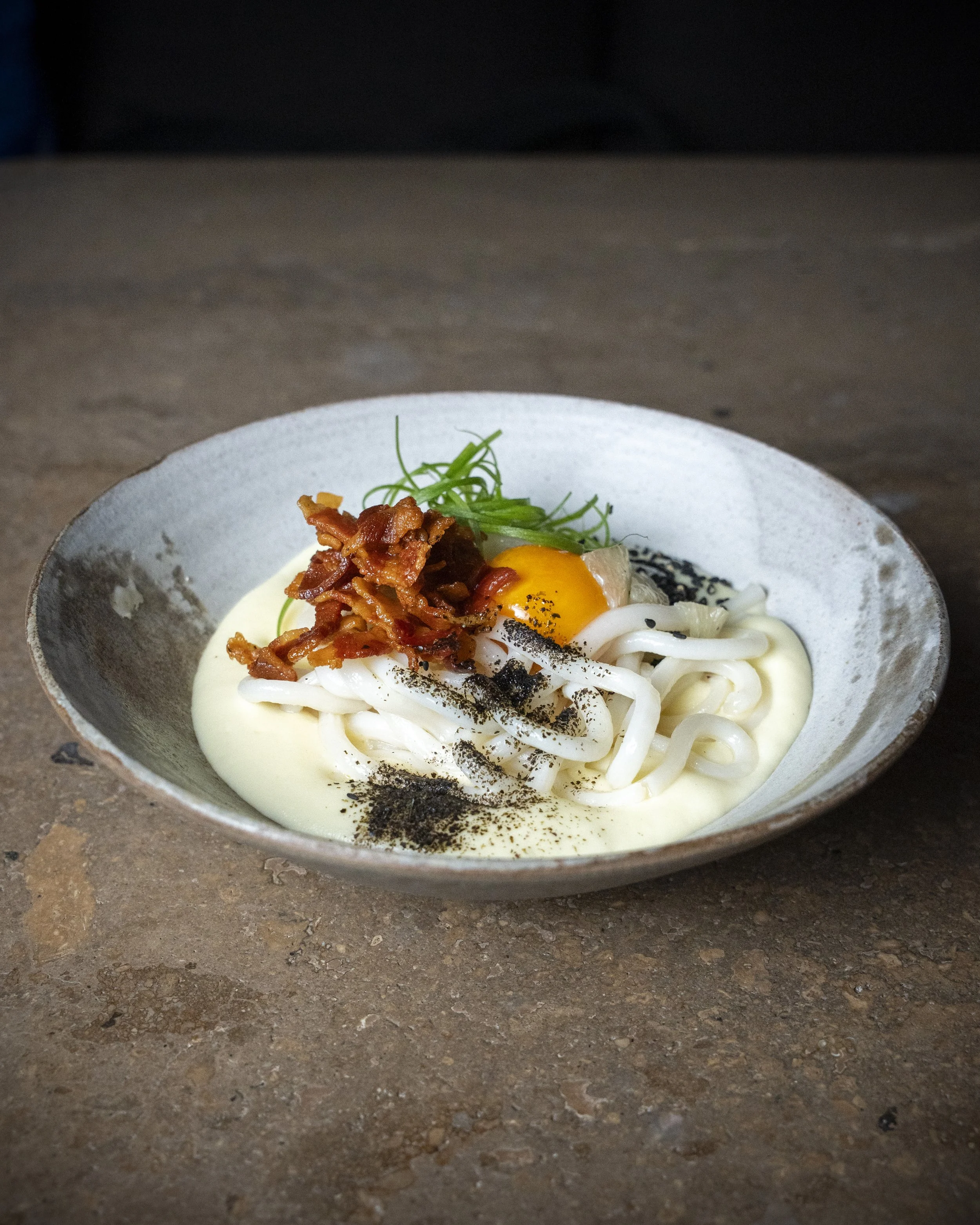 Bowl of Asian-style noodles with an egg, bacon, green onion, black pepper, and a creamy sauce on a rustic surface.