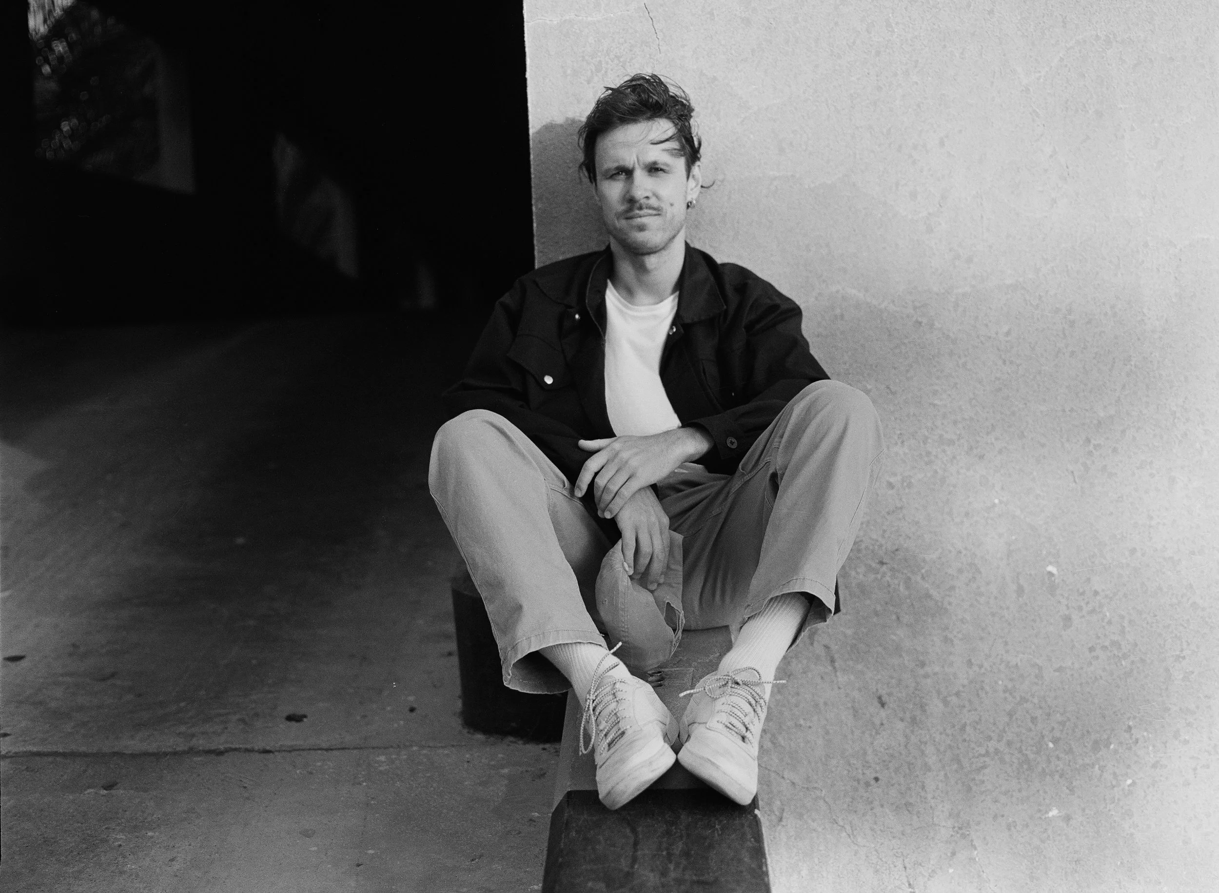 A young man with short hair and a mustache, sitting on a small stool with his legs crossed. He is wearing light-colored pants, a white T-shirt, and a dark jacket, and he is holding a cap in one hand. He is sitting against a textured wall, with a dark