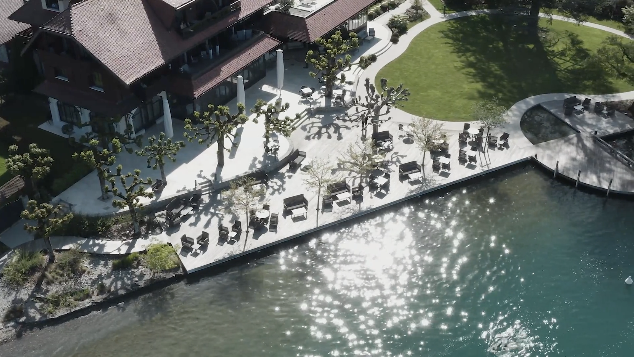 Aerial view of a waterfront outdoor patio with seating, trees, and a building, adjacent to a body of water with sunlight reflections.