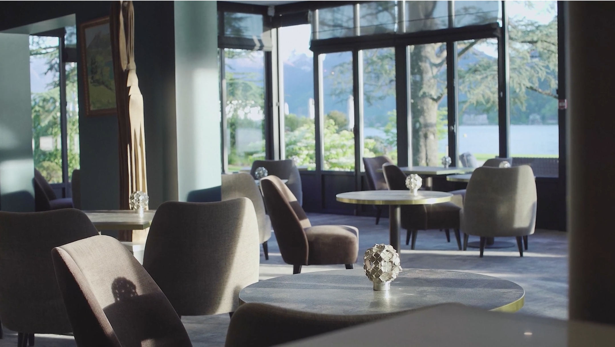 Interior of a restaurant or lounge with upholstered chairs and round tables, some decorated with small vases of white flowers, large windows with a view of greenery and water, and a mountain background.