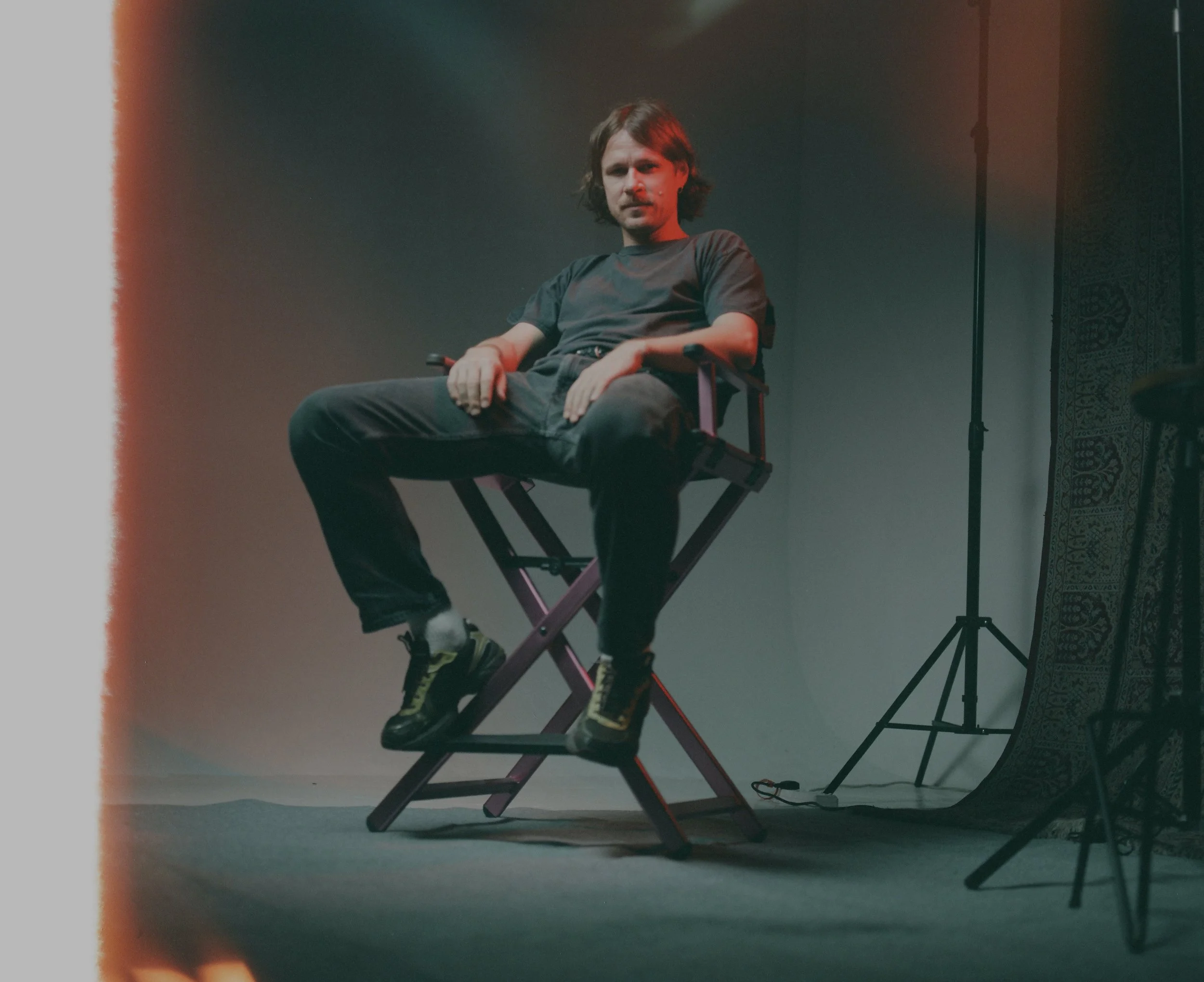 A man sitting on a director's chair in a studio setting with minimal equipment visible, including a light stand and a patterned curtain or backdrop to the side.