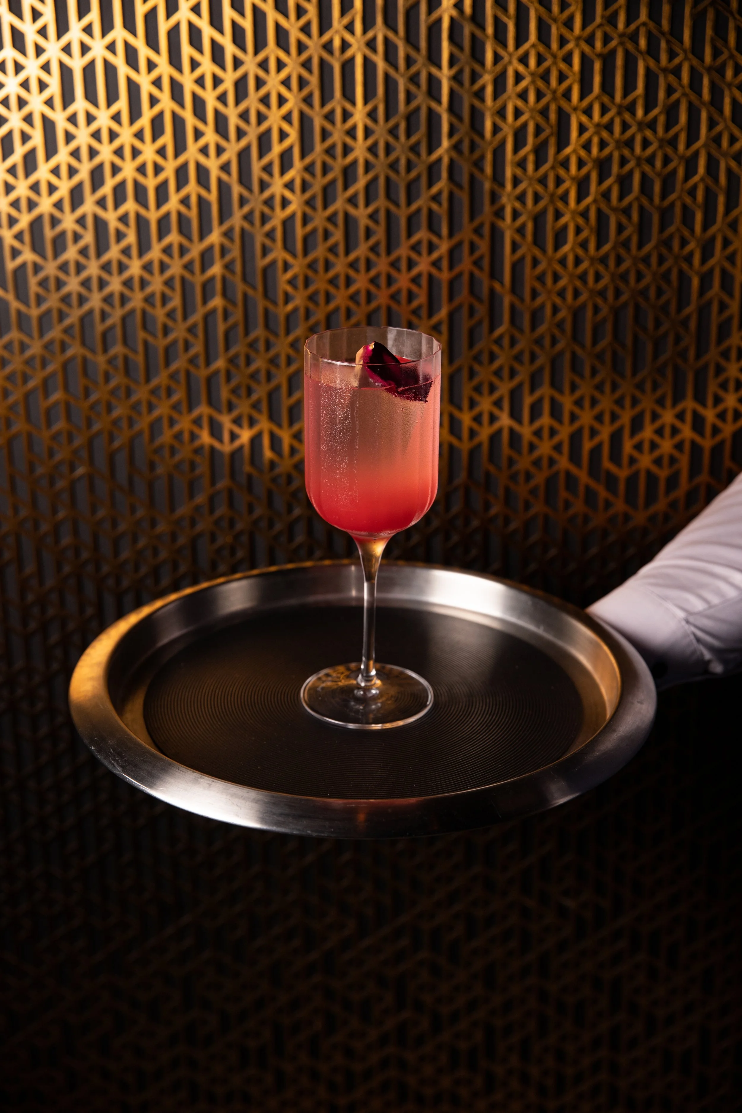 A pink cocktail with a dark cherry garnish on a silver tray, against a geometric patterned gold wall.