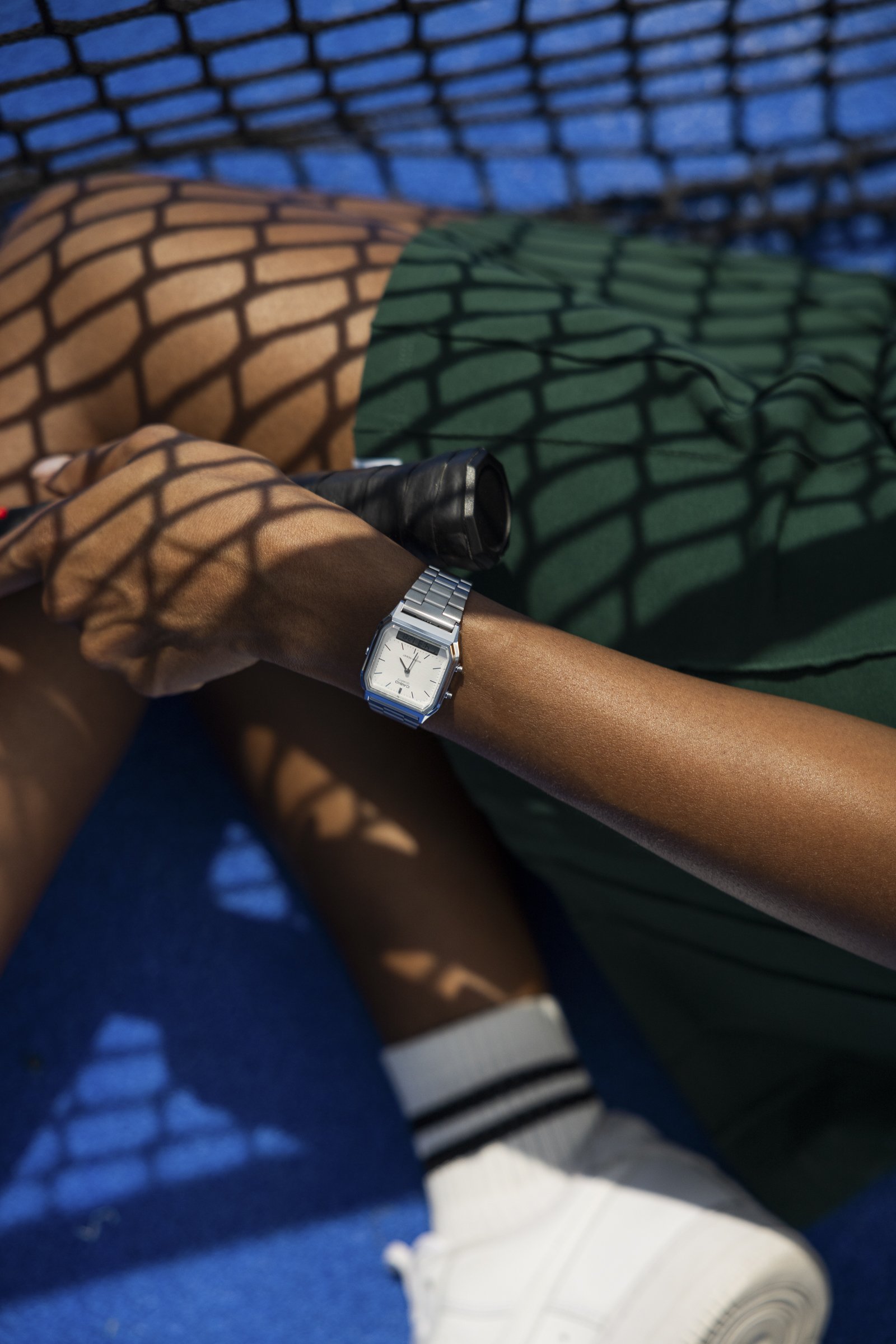 A person wearing a silver watch and white sneakers with black stripes, lying on a blue surface with a shadow pattern from a nearby fence.