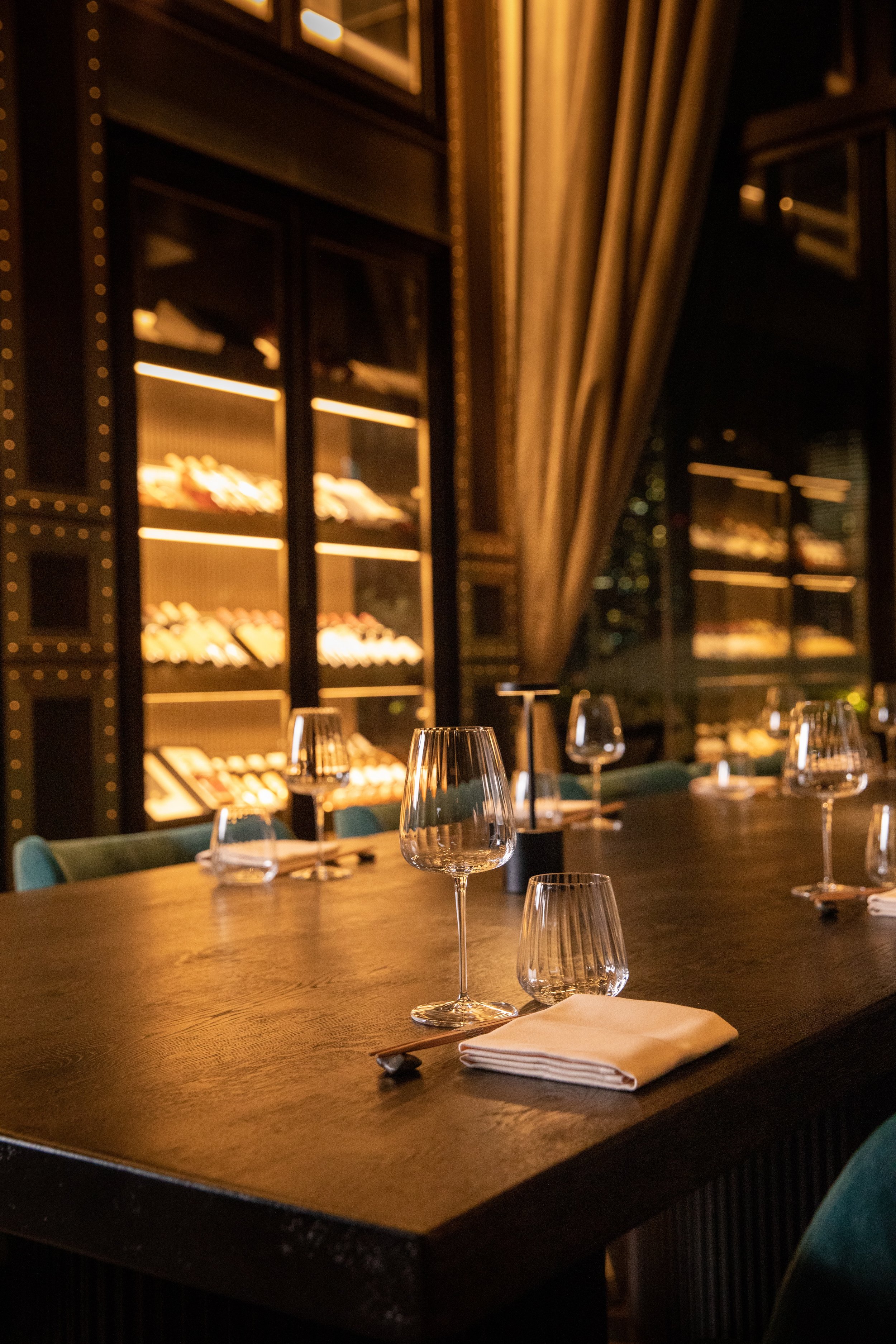 Elegant restaurant table set with wine glasses, napkins, and cutlery, with a wine display cabinet and a large curtain in the background.
