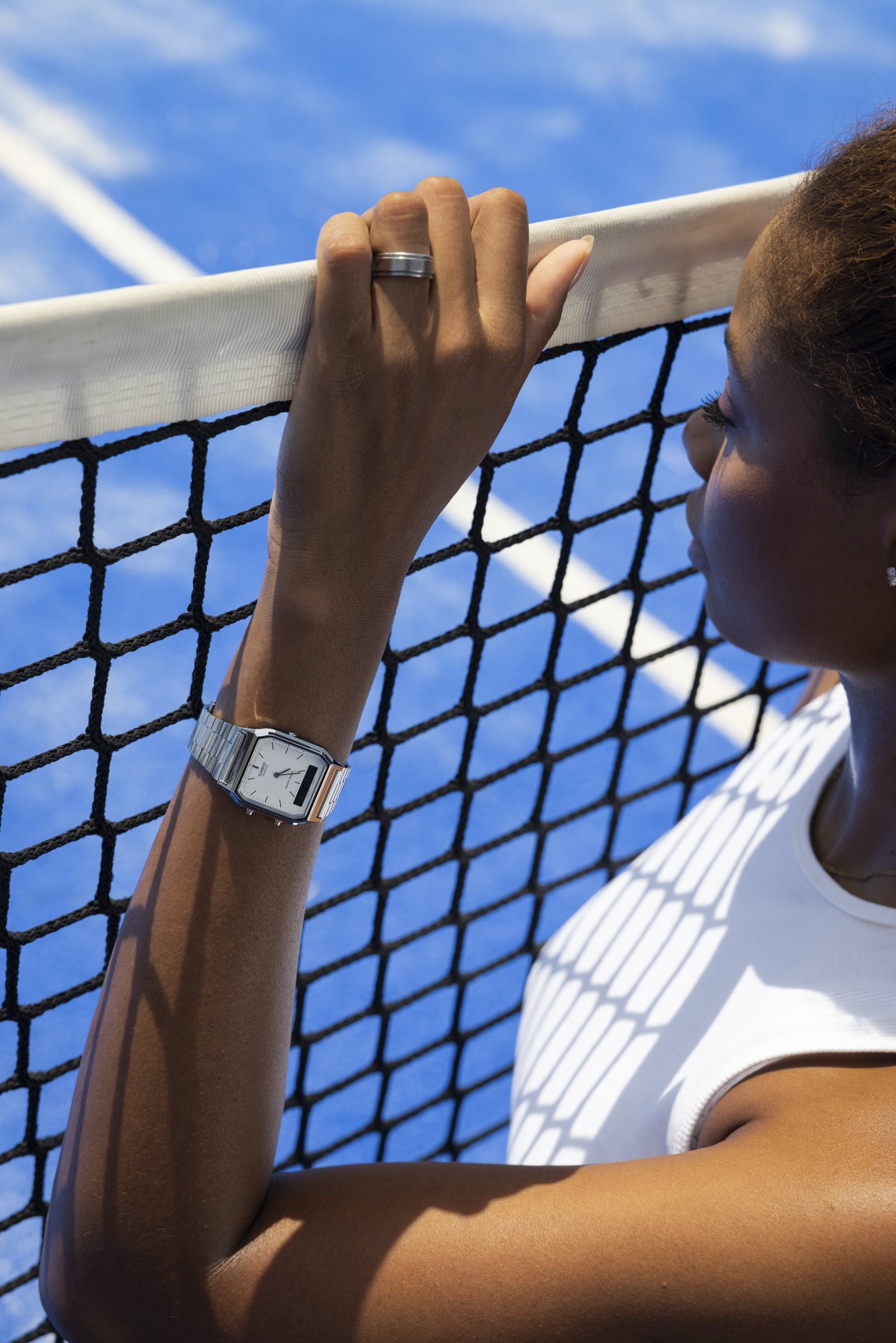 A woman wearing a silver watch and pearl earrings resting her arm on a tennis net, with a blue tennis court in the background.