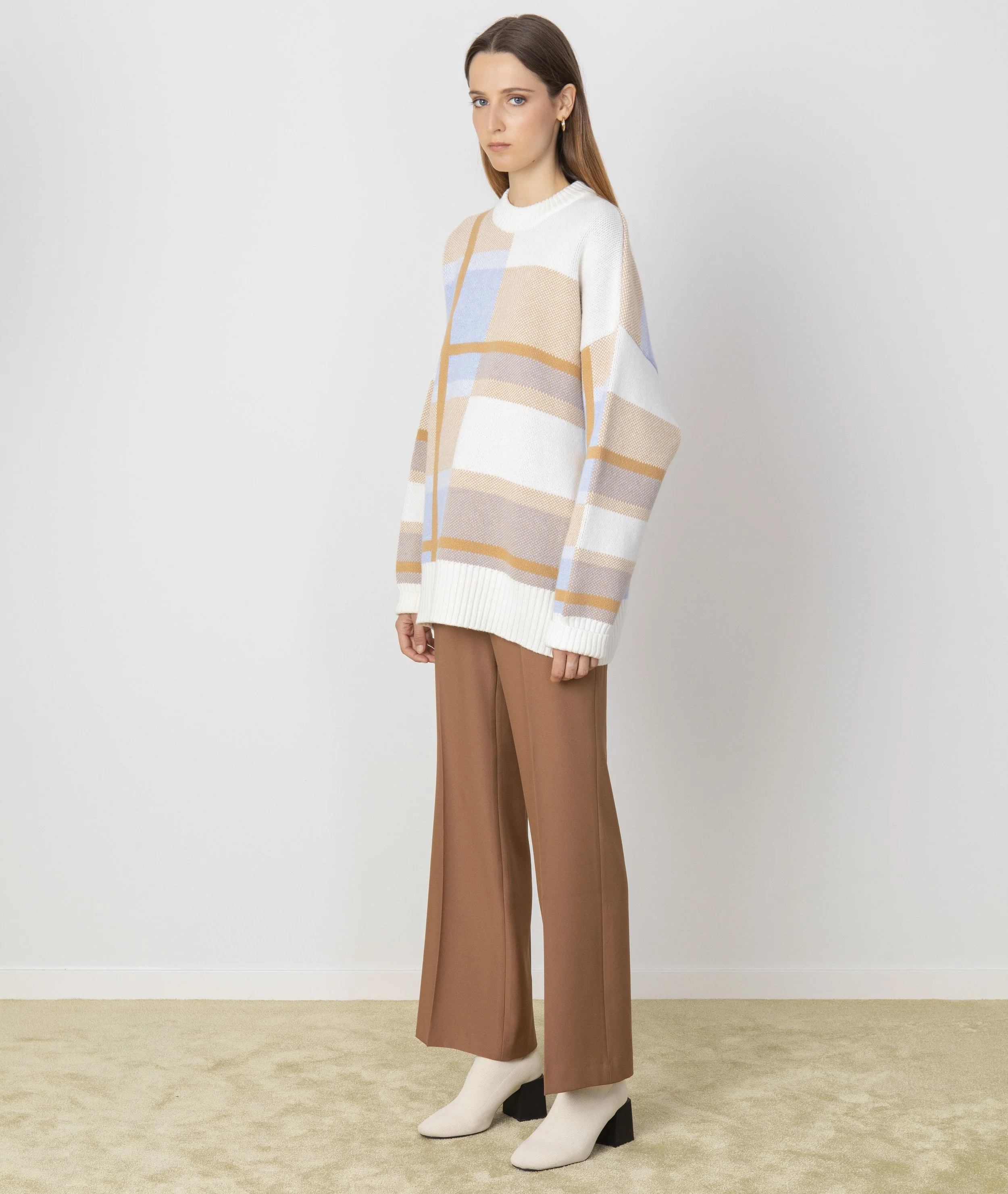 A woman standing in front of a plain white wall, wearing a long sleeve, oversized, checkered sweater in white, beige, blue, and tan colors, paired with brown wide-leg trousers and white ankle boots with black heel.