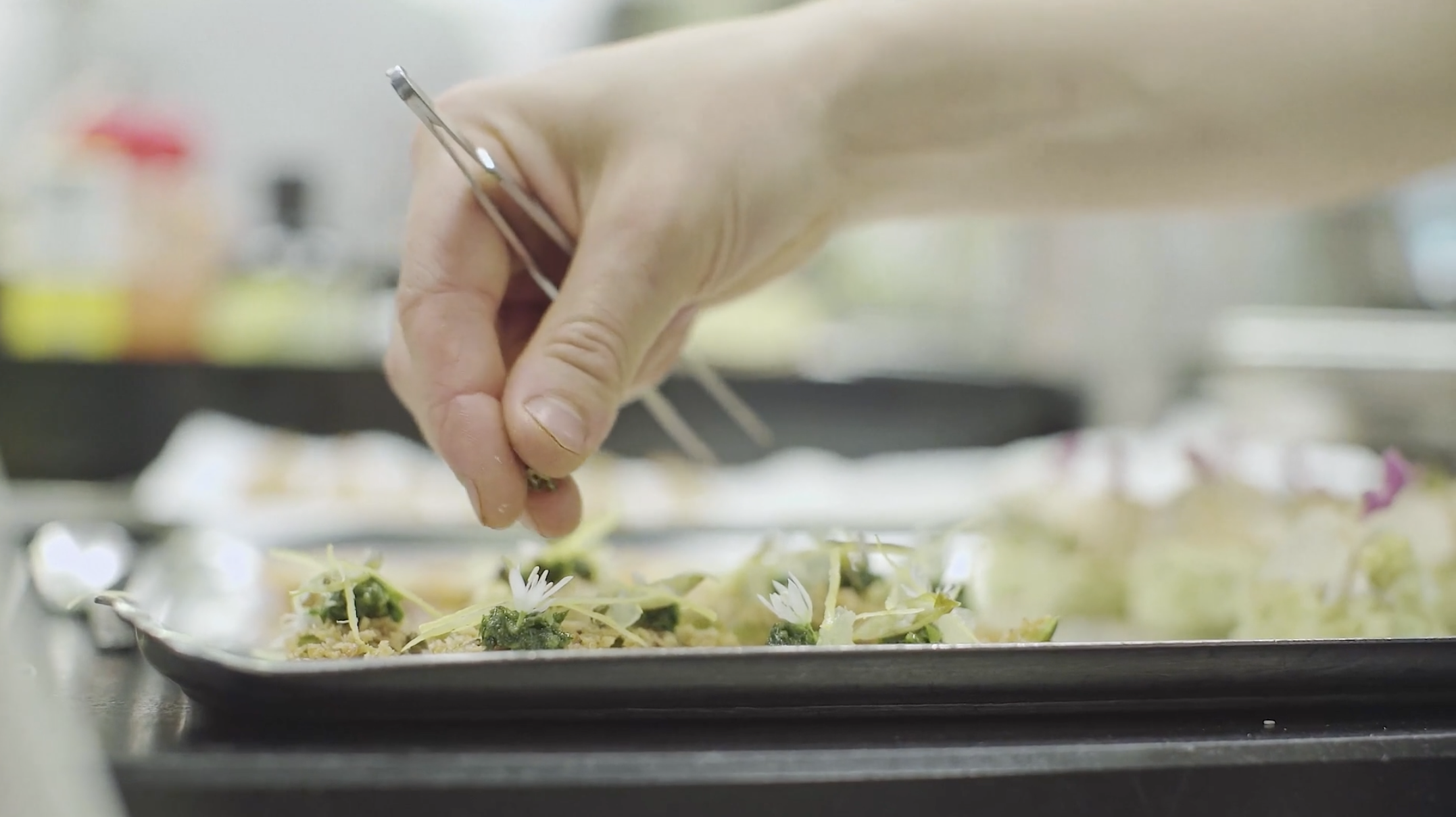 A chef's hand carefully decorates a gourmet dish with edible flowers and microgreens on a black tray in a professional kitchen.