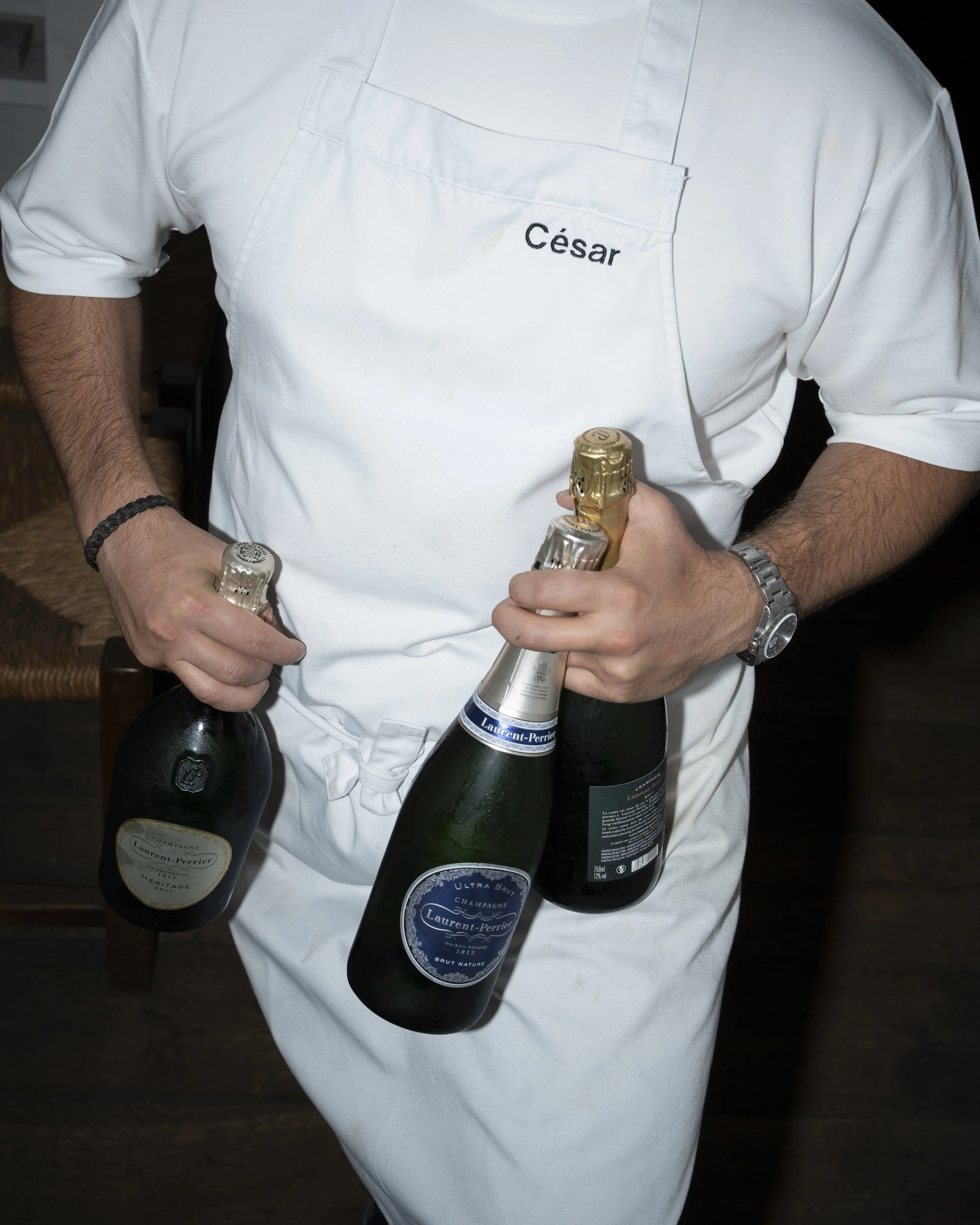 Person in a white uniform with the name 'César' holding three bottles of champagne.