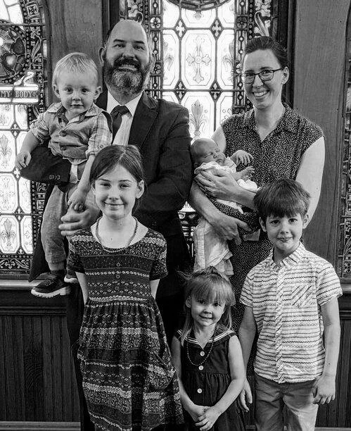 A black and white family portrait featuring two adults and five children standing in front of decorative stained glass windows. The man is holding a toddler and the woman is holding an infant. Three other children stand in front, smiling at the camera.