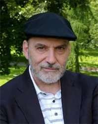 Man wearing a black flat cap, white polo shirt, and dark blazer standing outdoors with greenery in the background.