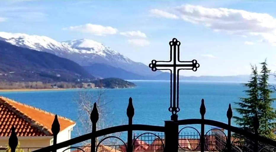 View of a lake with a metal cross on a gate in the foreground, snow-capped mountains in the background, and trees on the right.