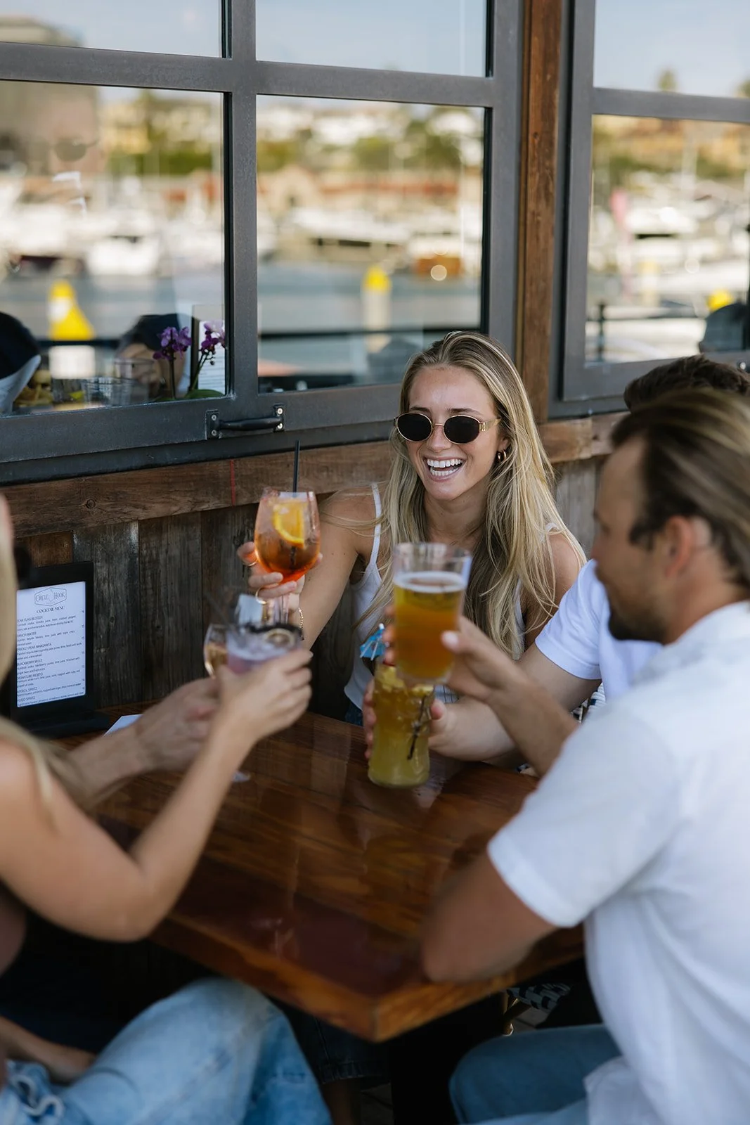 A group of friends enjoying drinks at a restaurant with windows overlooking a marina with boats and water.