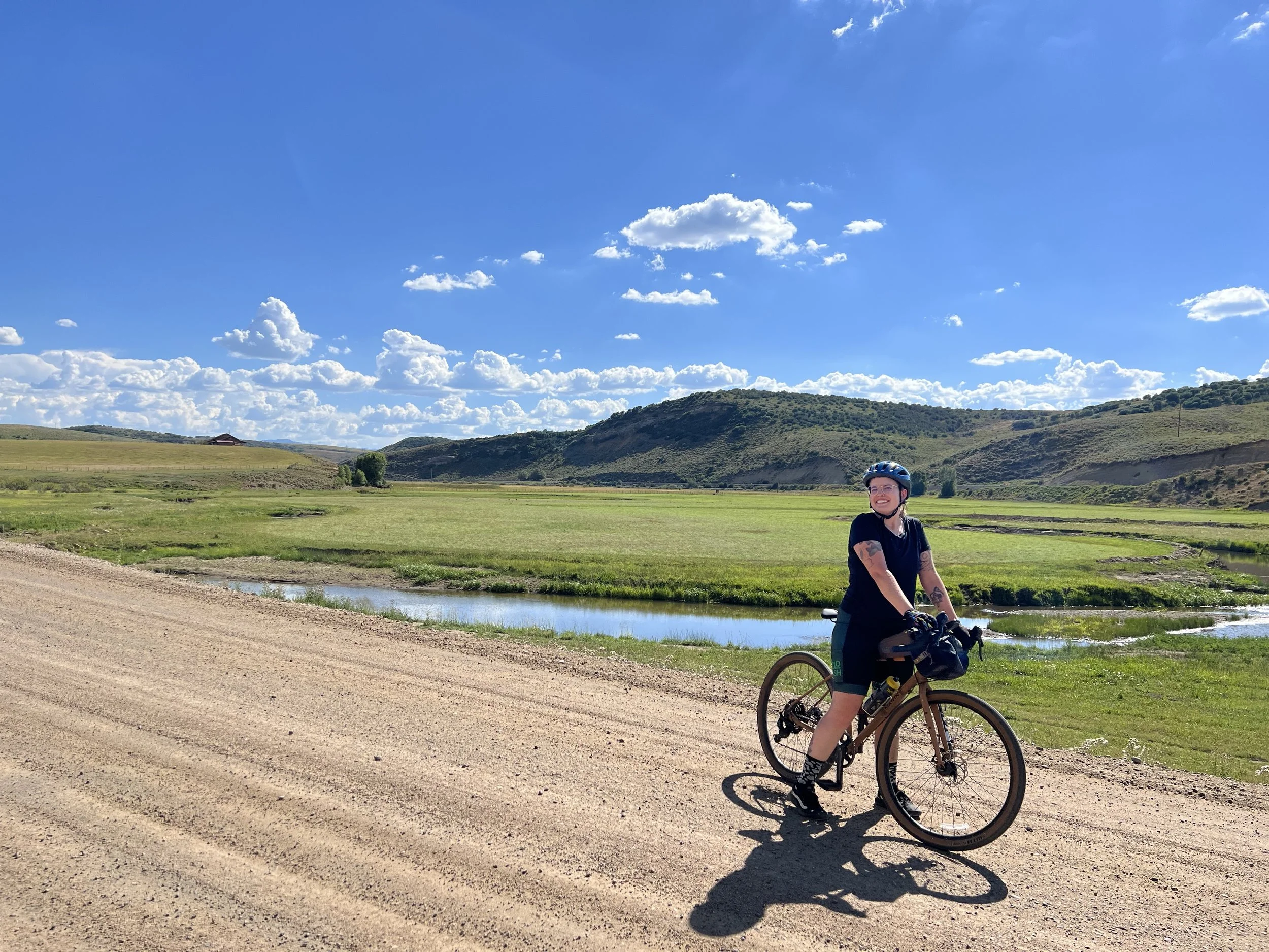 A white person is standing over their bicycle on an dirt road. In the foreground is a field with a stream running through it. In the background are low, rolling foothills with trees placed sporadically. There is a clear blue sky with fluffy clouds.