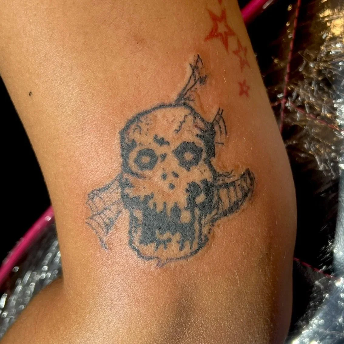 Tattoo of a skull with spider web in the eye socket and an eye on its cheek, outlined in black on a person's skin near the thigh.