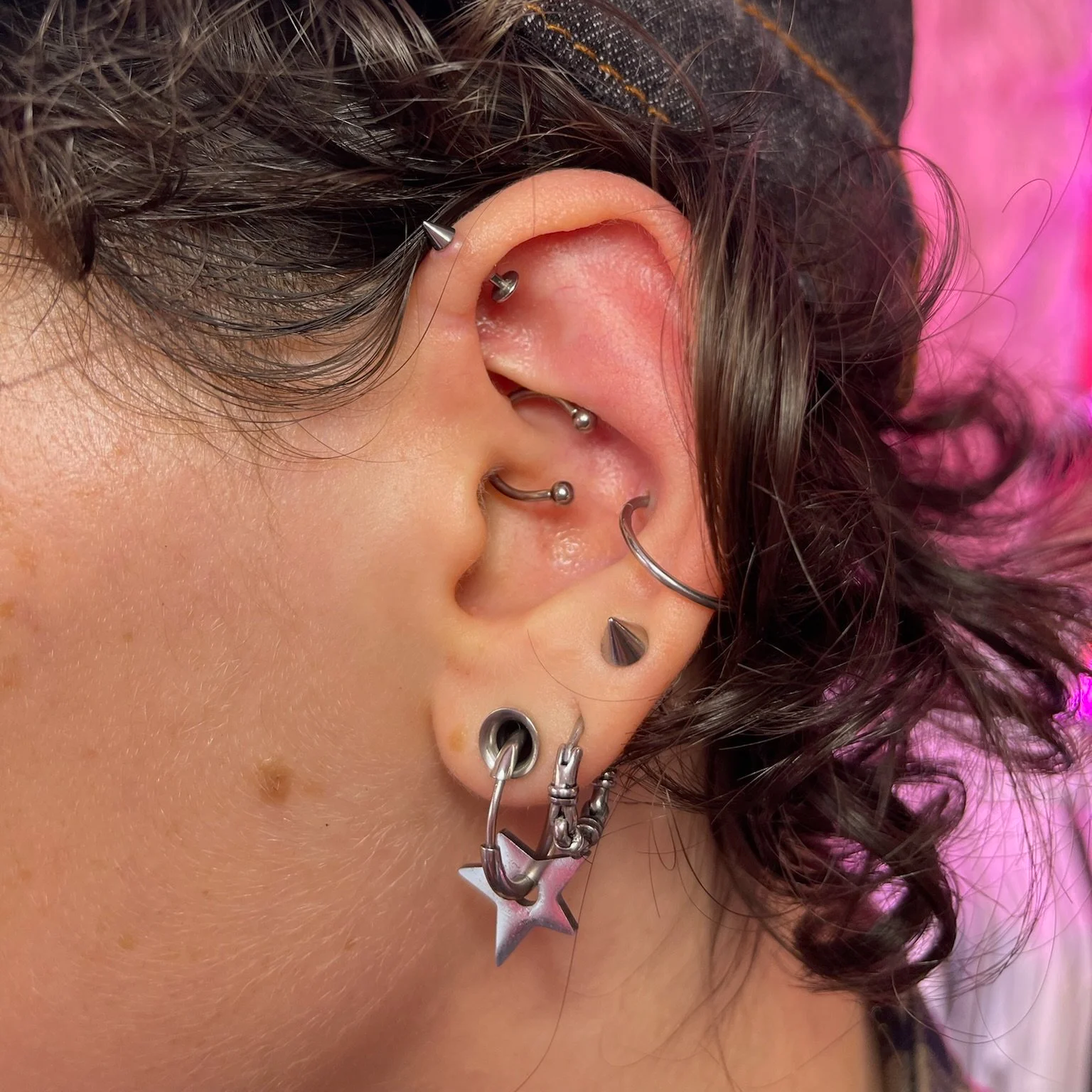 Close-up of a person's ear with multiple silver earrings, including a star-shaped earring, a hoop, and various studs, against a pink background.
