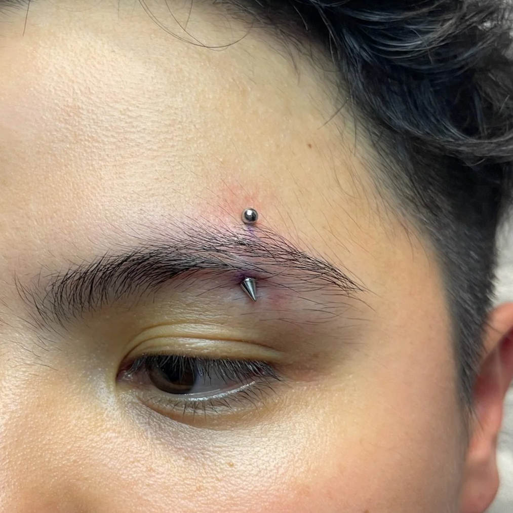 Close-up of a person's eye with a eyebrow piercing and a vertical labret piercing above the eye.