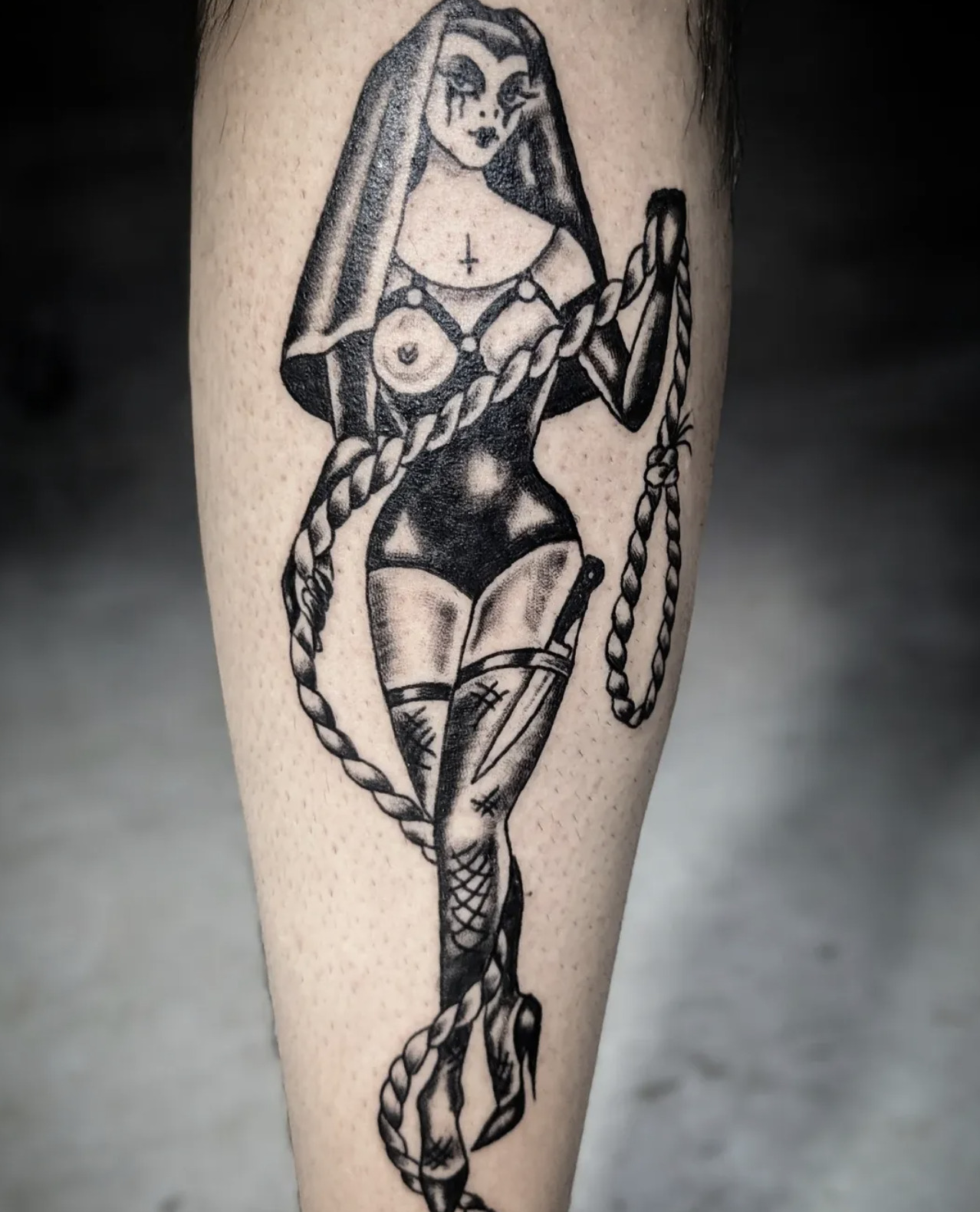 Black and gray tattoo of a woman with Dolly Parton style hair, tattoos on her chest, and stockings, holding a whip, with a confident expression.