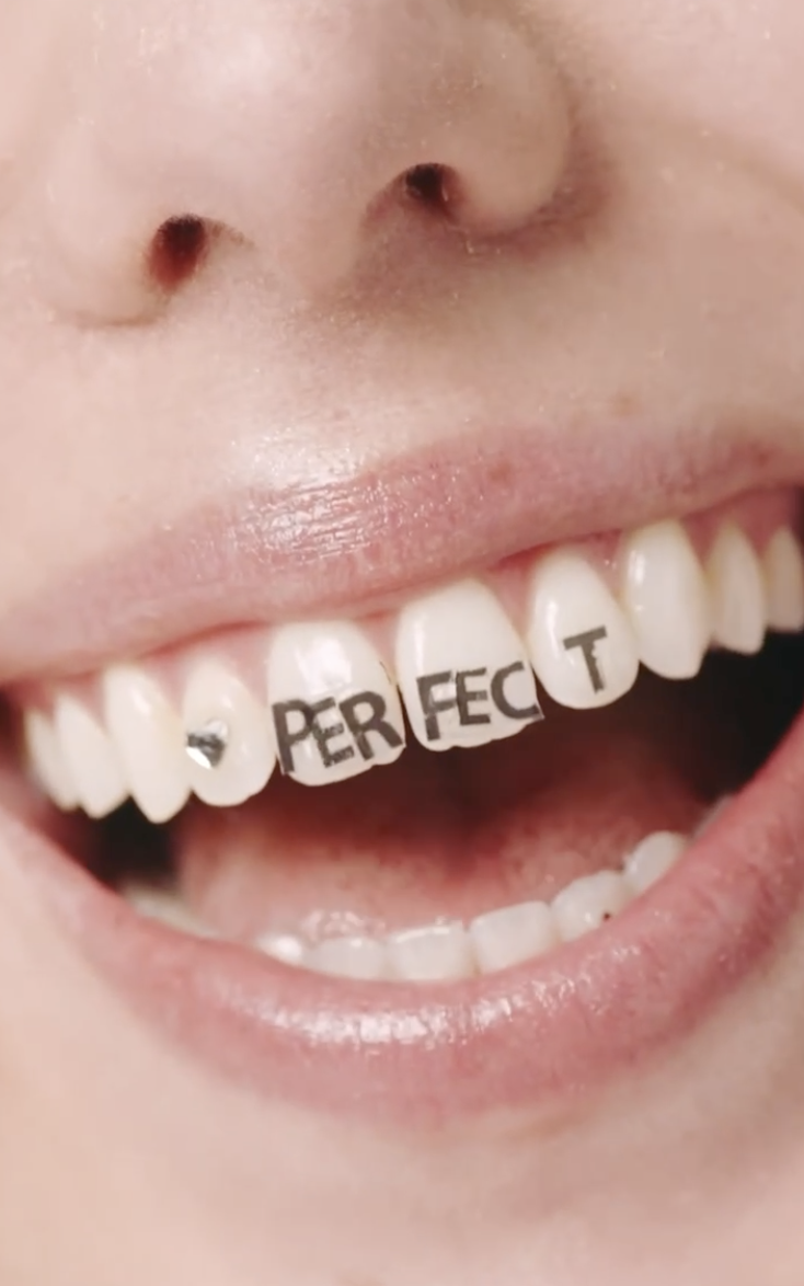 Close-up of a person's mouth showing gold teeth spelling out 'FAME' with tongue and lips slightly open.