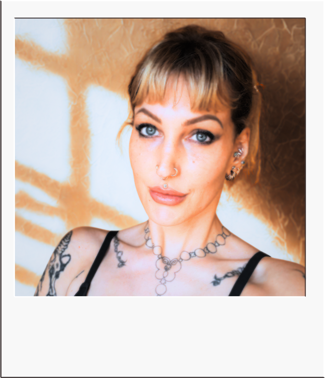 A woman with blonde hair and blue eyes taking a selfie against a pink background, wearing a black top with red accents and tattoos on her arms.
