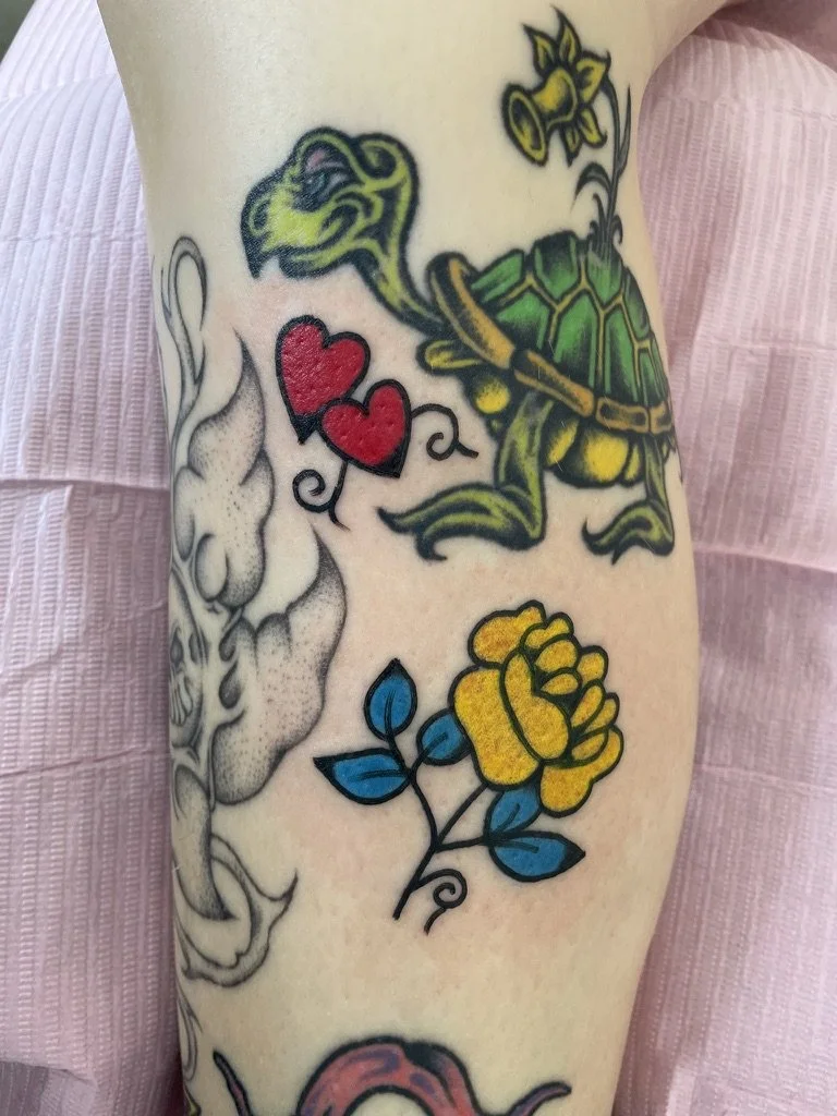 Tattoo of a turtle with a dragon-like head, two red hearts, a yellow flower with green leaves, and a pink laugh character on person's arm.