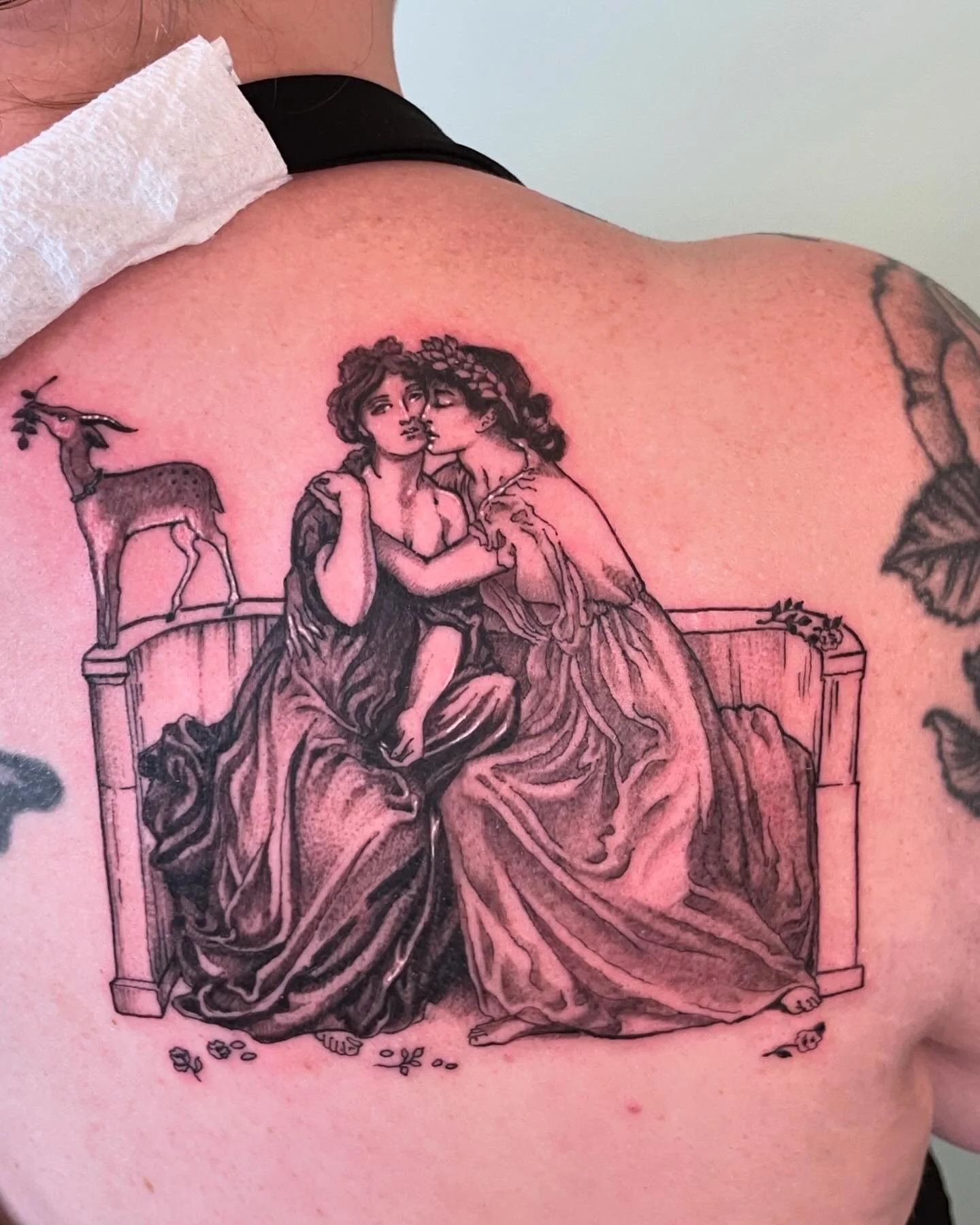 Tattoo of two women sitting on a bench, with one resting her head on the other's shoulder, and a deer on the left side of the bench.