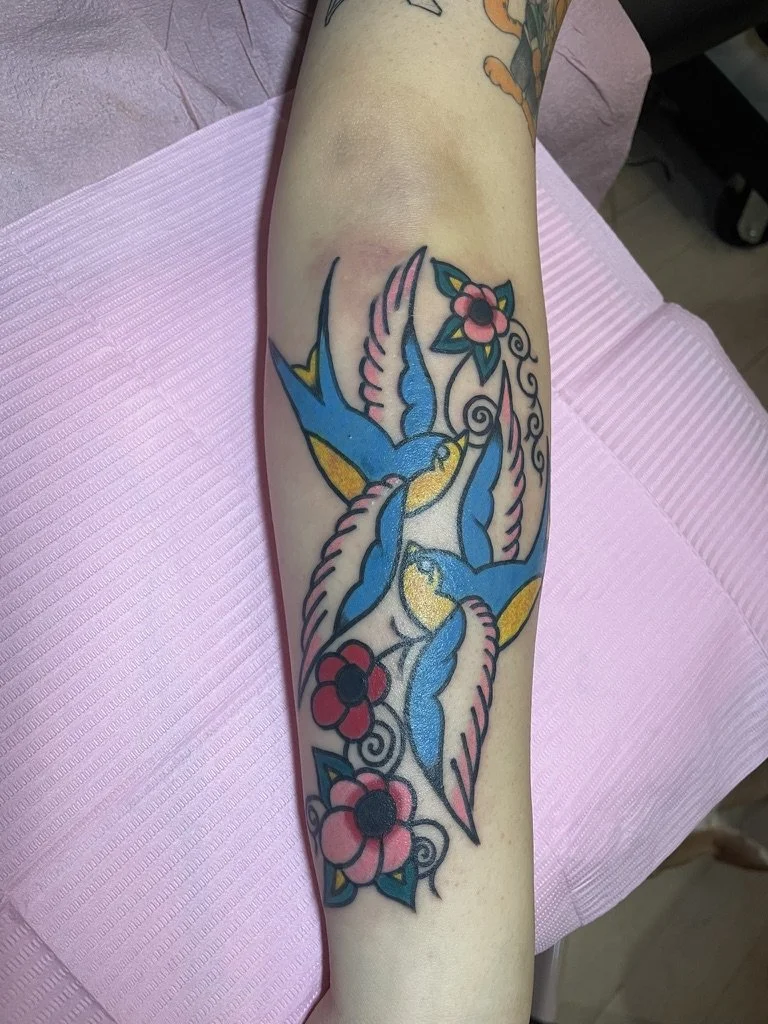 Tattoo of three blue and yellow hummingbirds flying around pink and red flowers with green leaves on a person's forearm.