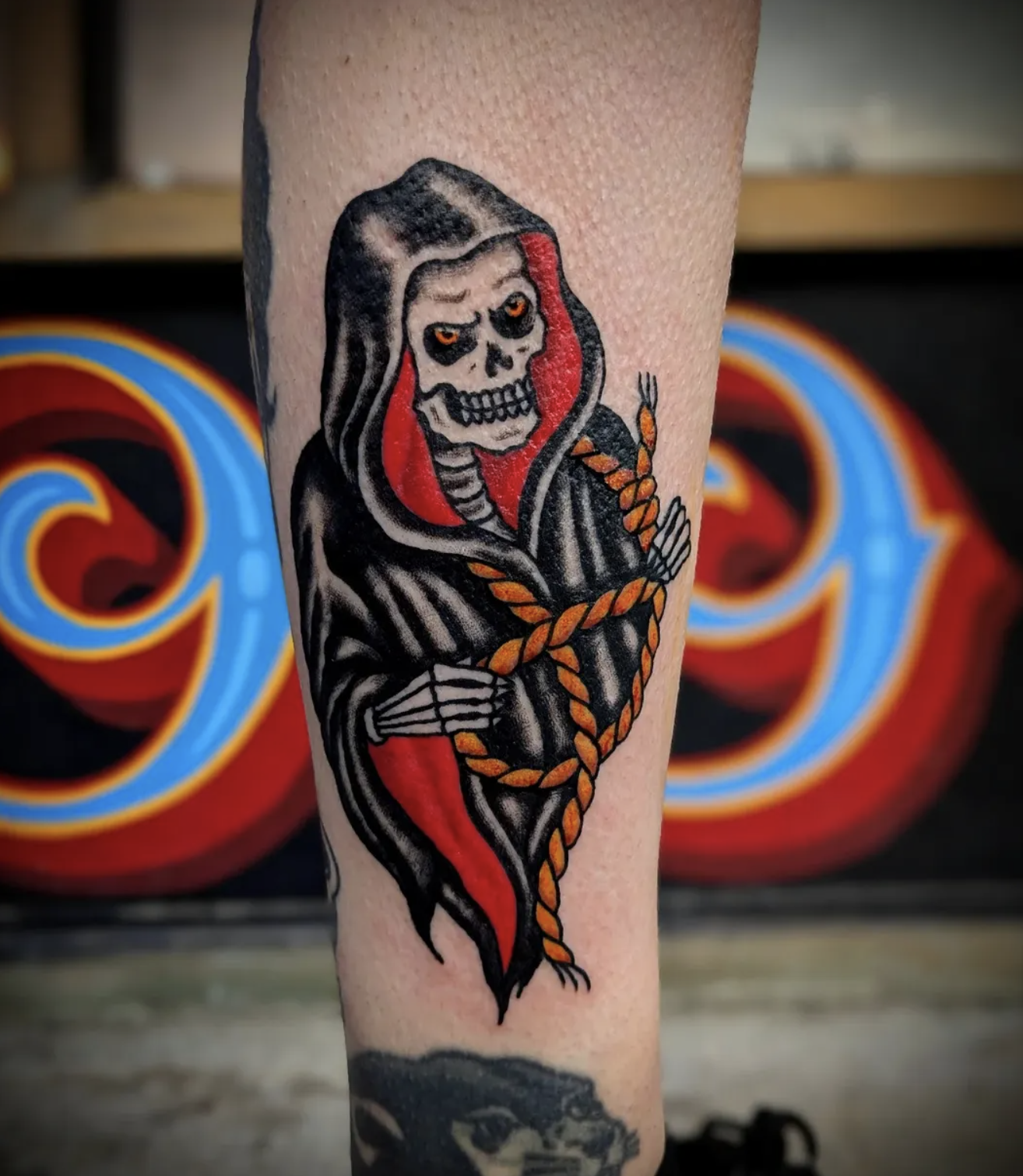 Tattoo of a skeleton in a hooded cloak with red accents, crossing its arms with a rope around its shoulders.