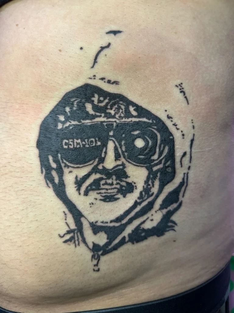 Tattoo of a man in sunglasses with a mustache and beard, wearing a hoodie, on skin.
