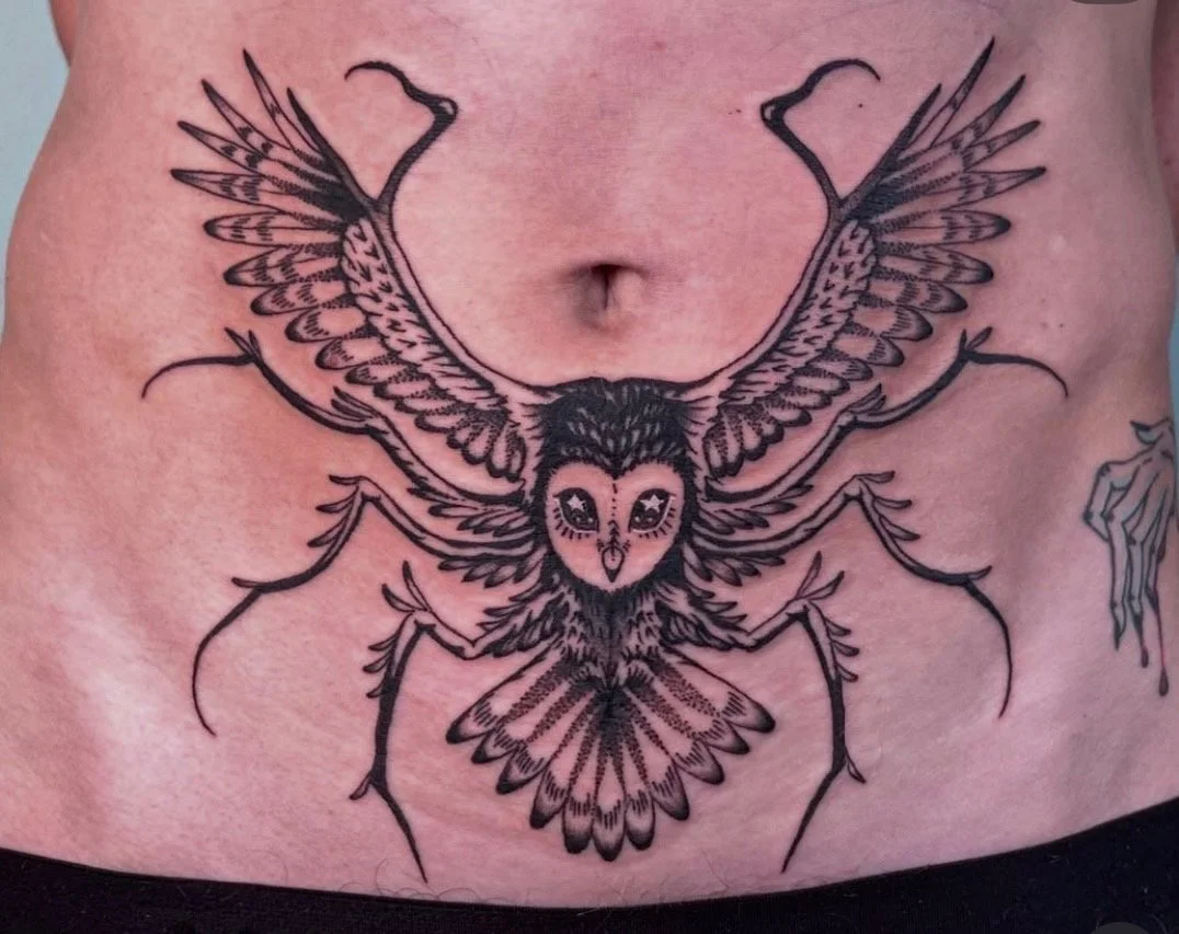 A tattoo on a person's abdomen depicts a detailed black line drawing of an owl with outstretched wings, combined with a spider's body and legs.