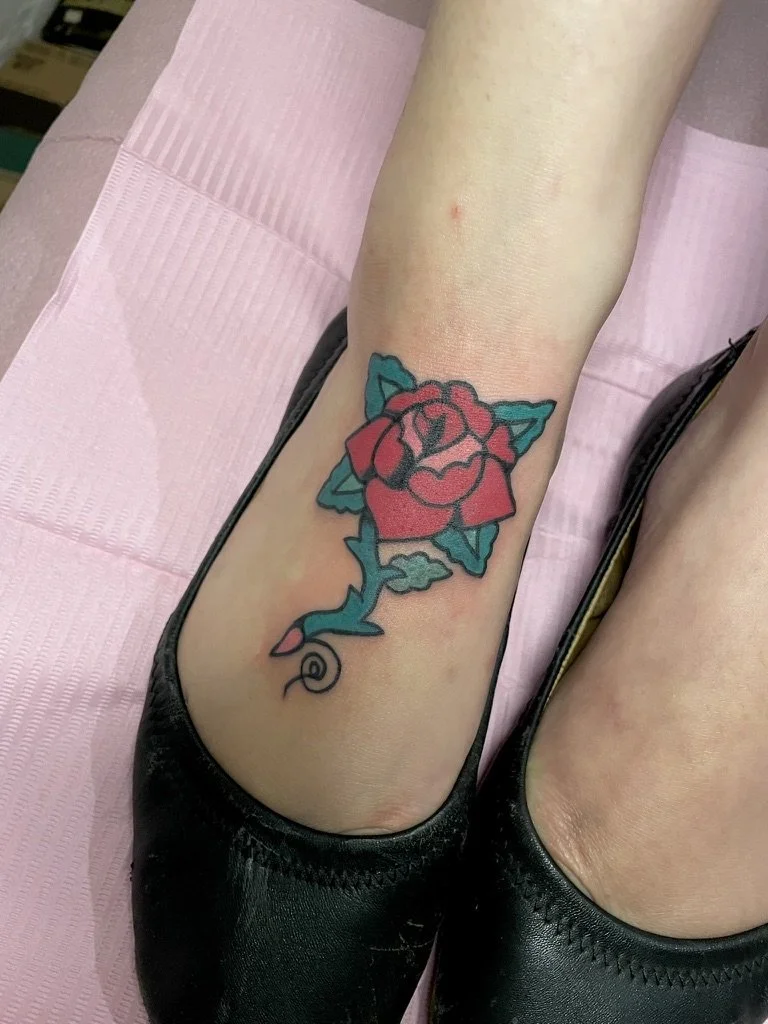 Tattoo of a cartoonish rose with leaves on an ankle, in pink, green, and black ink.