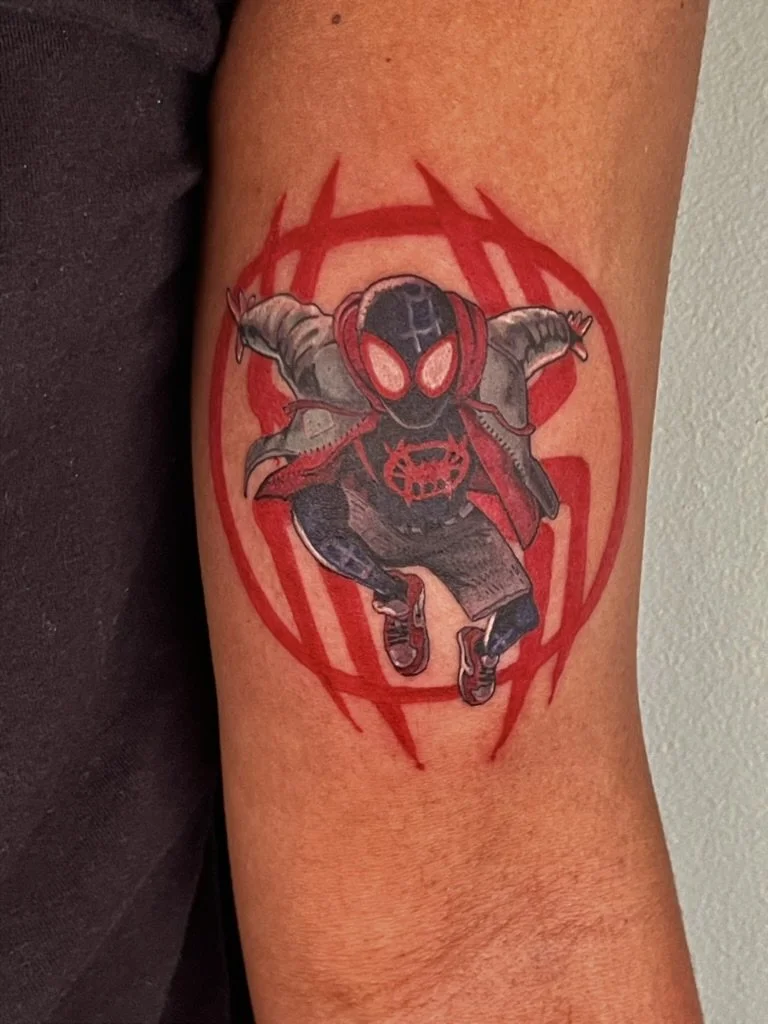 A tattoo of Spider-Man in a black costume with red accents, crouching with arms outstretched, inside a red heart outline. The tattoo features bold black, red, and gray ink.