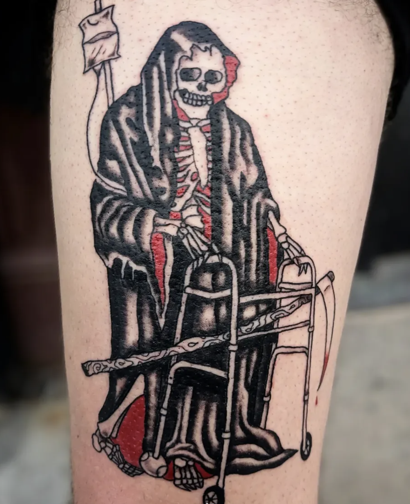 Tattoo of a skeleton in a hooded cloak sitting in a wheelchair, holding its ribs with one hand and a scythe in the other. The skeleton has a red line on its face and body, and a small flag on a pole is behind it.