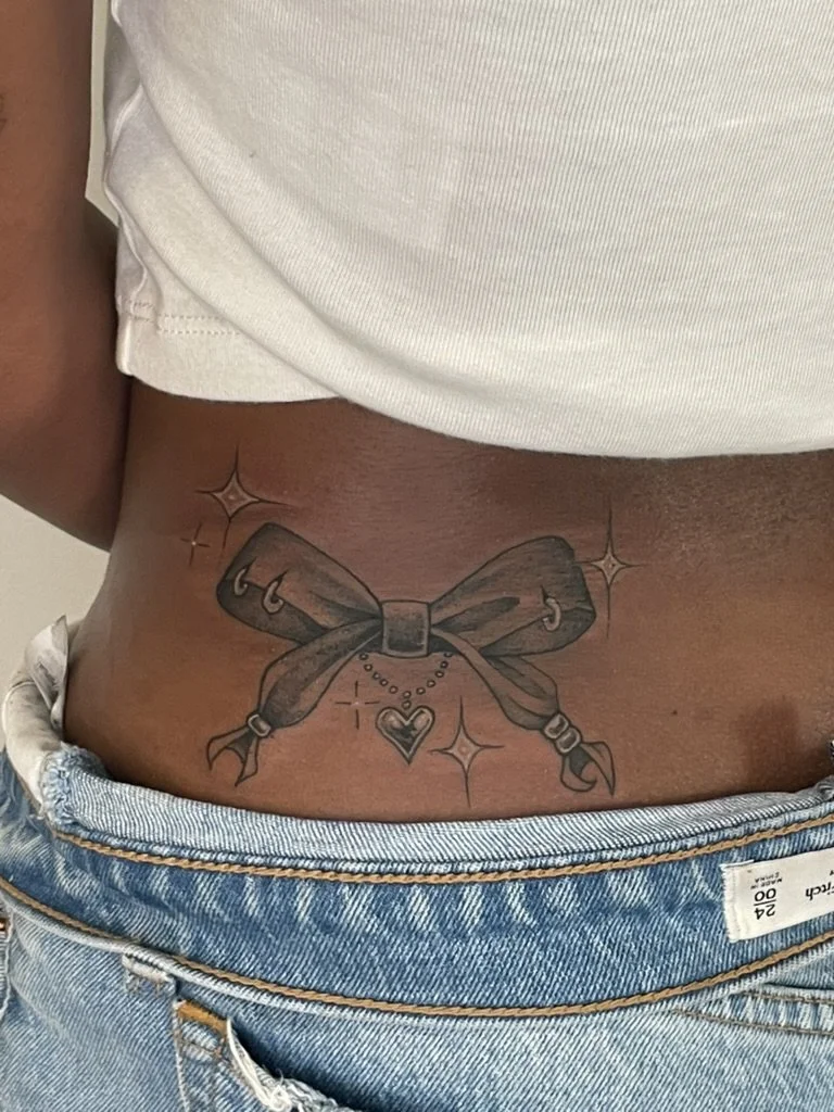 Tattoo of a large bow with a heart charm hanging from it, surrounded by small stars and sparkles, on the person's lower abdomen above the jeans.