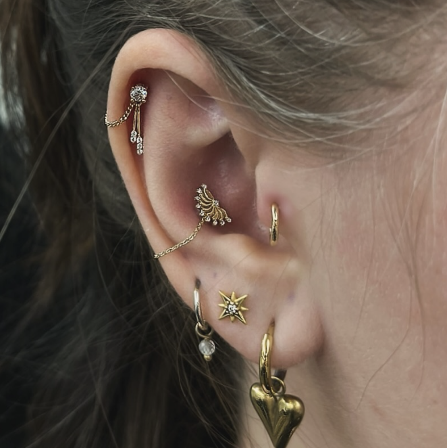 Close-up of a person's ear adorned with multiple gold and silver earrings, including a star-shaped earring, a heart-shaped earring, and a chain connected to various earrings, with light brown hair partially covering the ear.