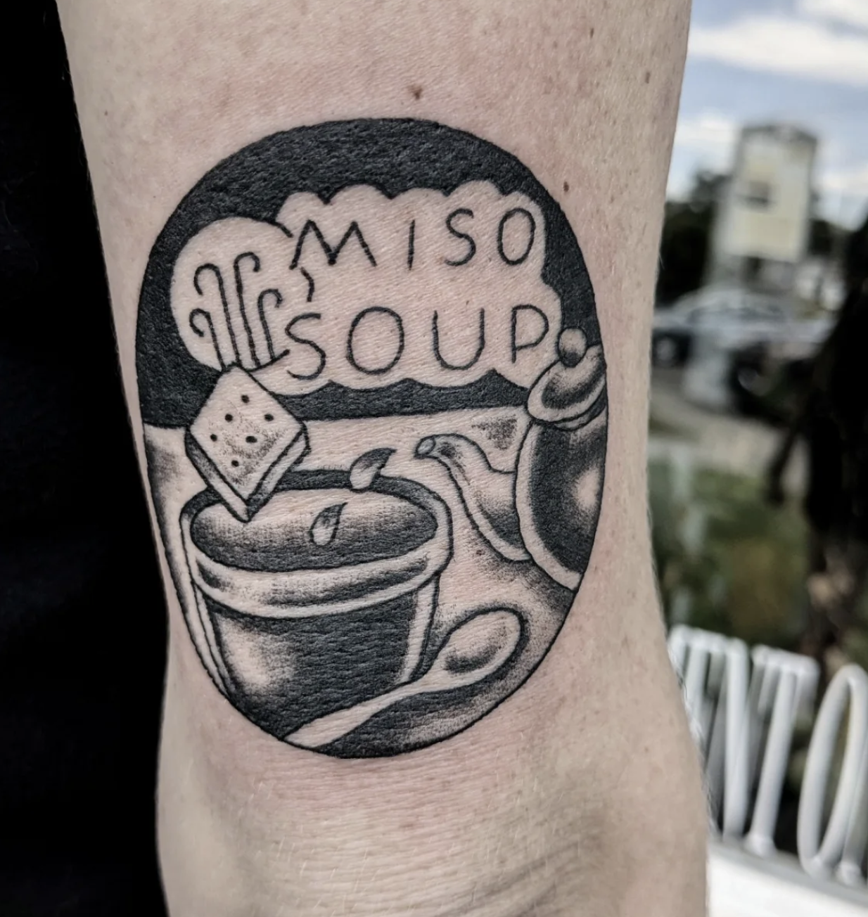 Tattoo of a coffee cup with a spoon, a slice of cake, and a teapot inside a circular frame, with the text 'I MISOUP.'